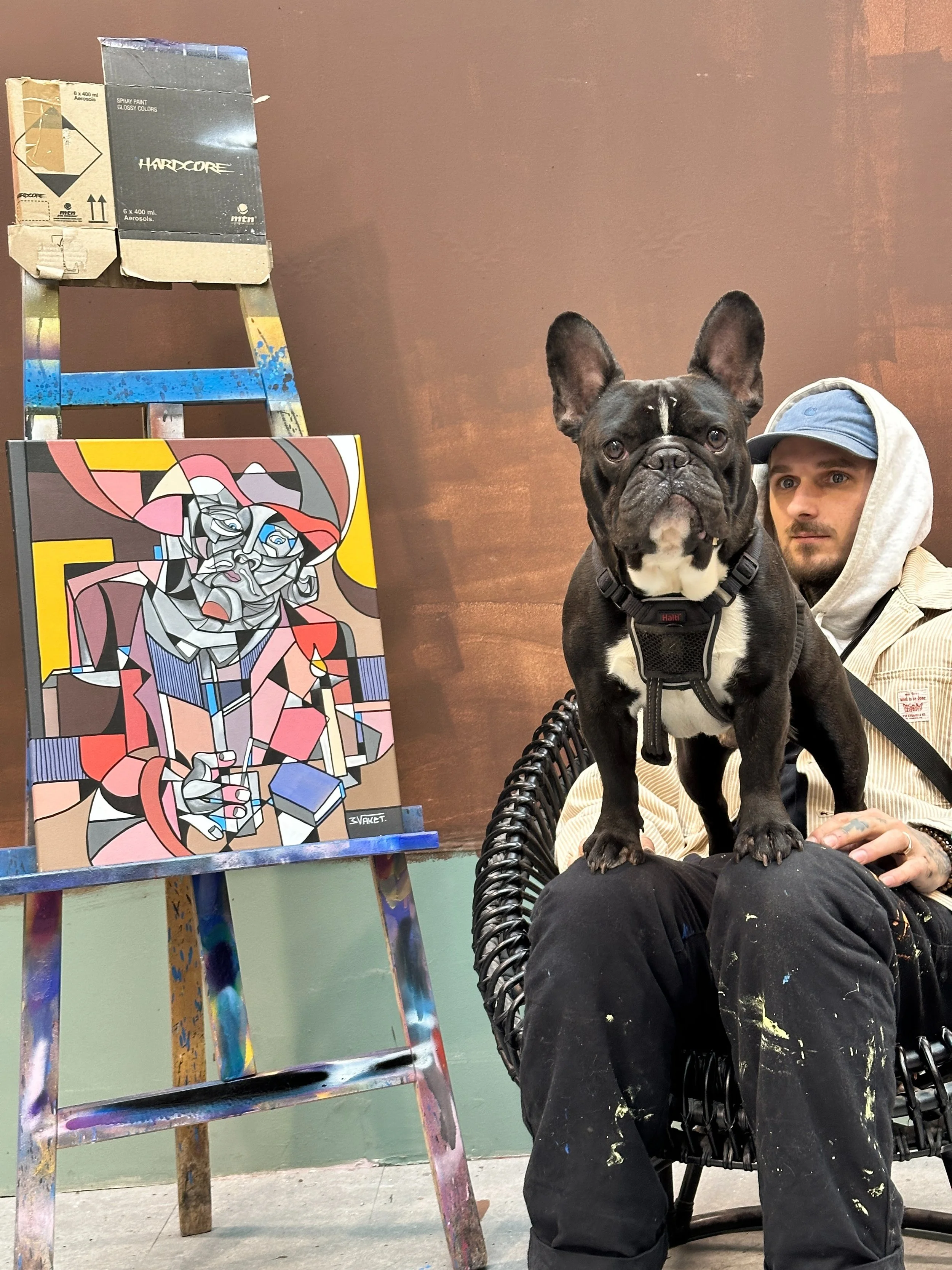 Un homme assis dans une chaise en osier, portant un hoodie et un chapeau, avec un bouledogue français debout sur ses genoux. À côté, il y a une peinture de style cubiste d'une femme portant un chapeau, placée sur une échelle recouverte de peinture