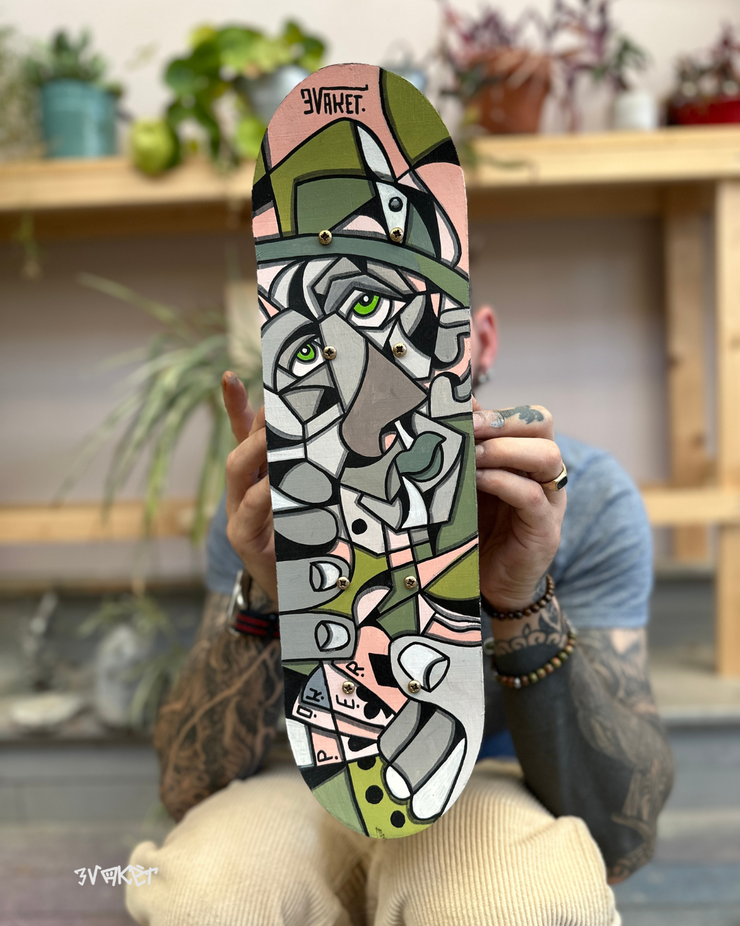 Planche à roulettes avec une illustration de style cubisme représentant une femme avec un chapeau gris et des yeux verts, tenant un objet. Personne tenant la planche avec des tatouages et des bracelets visible sur les bras. Arrière-plan avec des plantes en pot et bibliothèque en bois.