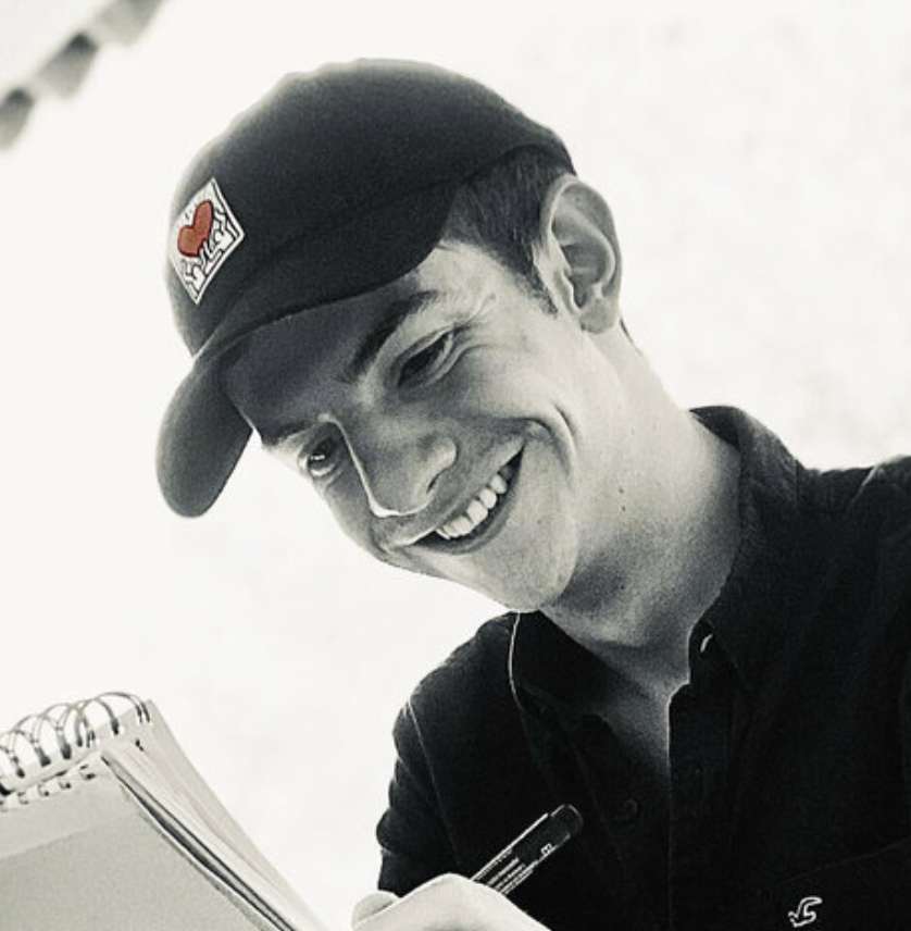 Jeune homme souriant portant une casquette noire avec un logo rouge, écrivant dans un carnet, avec des écouteurs dans les oreilles.