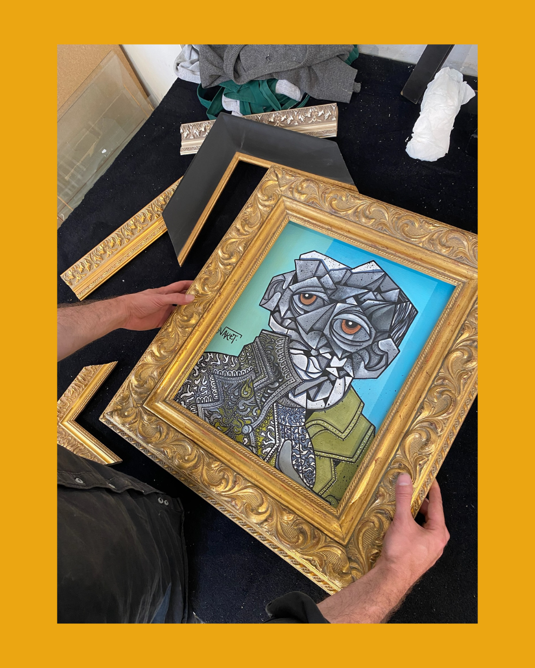 Une personne tient un tableau avec un portrait artistique d'un animal anthropomorphe, entouré de cadres dorés. Le tableau utilise des formes géométriques et un style vibrant.