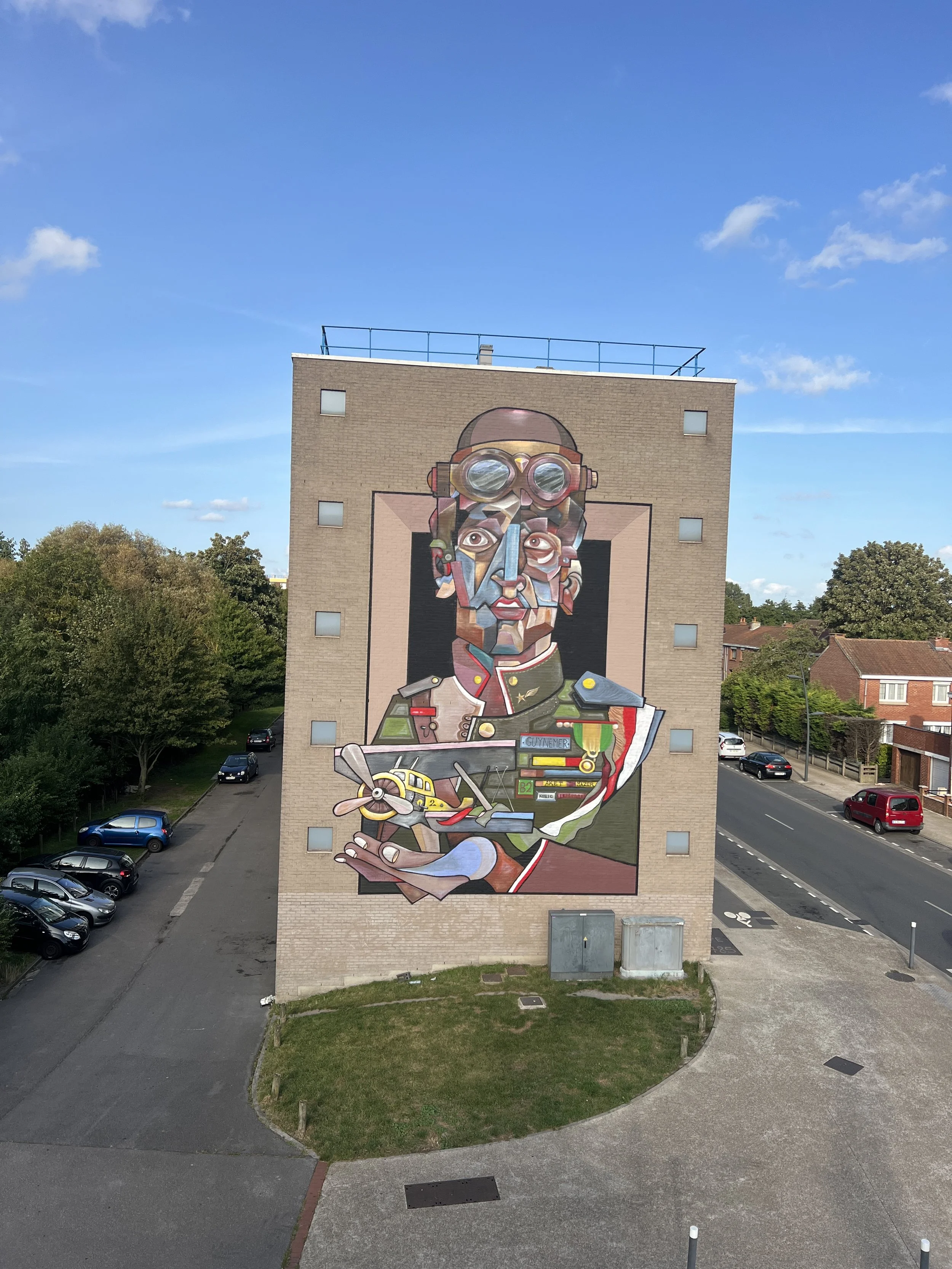 Murale artistique représentant un homme en uniforme militaire avec des lunettes de plongée sur la tête, tenant un modèle d'avion dans la main. La fresque est colorée et géométrique. Cela est peint sur la façade de dunkerque; street art
