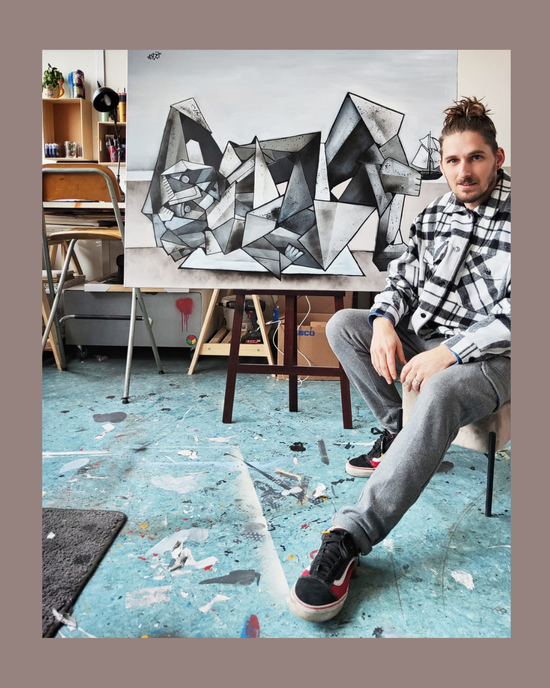 Un homme assis côte à côte avec une peinture abstraite géométrique représentant un animal, probablement un chat, dans un atelier d'art. L'atelier contient des étagères, des chaises, et un sol taché de peinture.