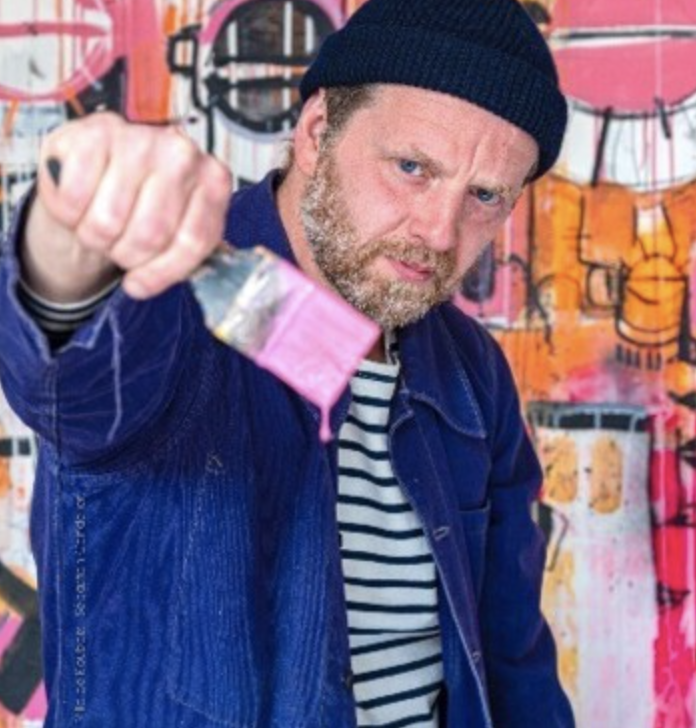 Un homme portant un bonnet noir et une veste bleue, tenant une bombe de peinture en spray, avec un mur graffiti coloré en arrière-plan.
