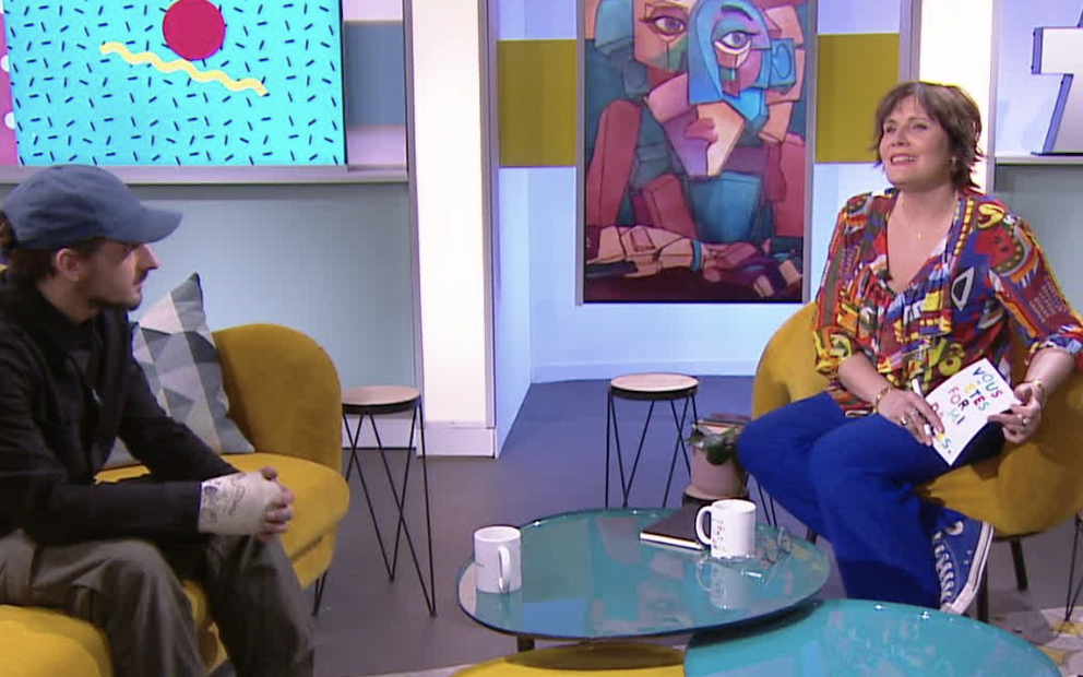 Une femme et un homme assis dans un salon de télévision, avec un écran d'ordinateur affichant une animation colorée en arrière-plan. La femme tient un livre, et il y a deux tasses sur la table en verre devant eux.