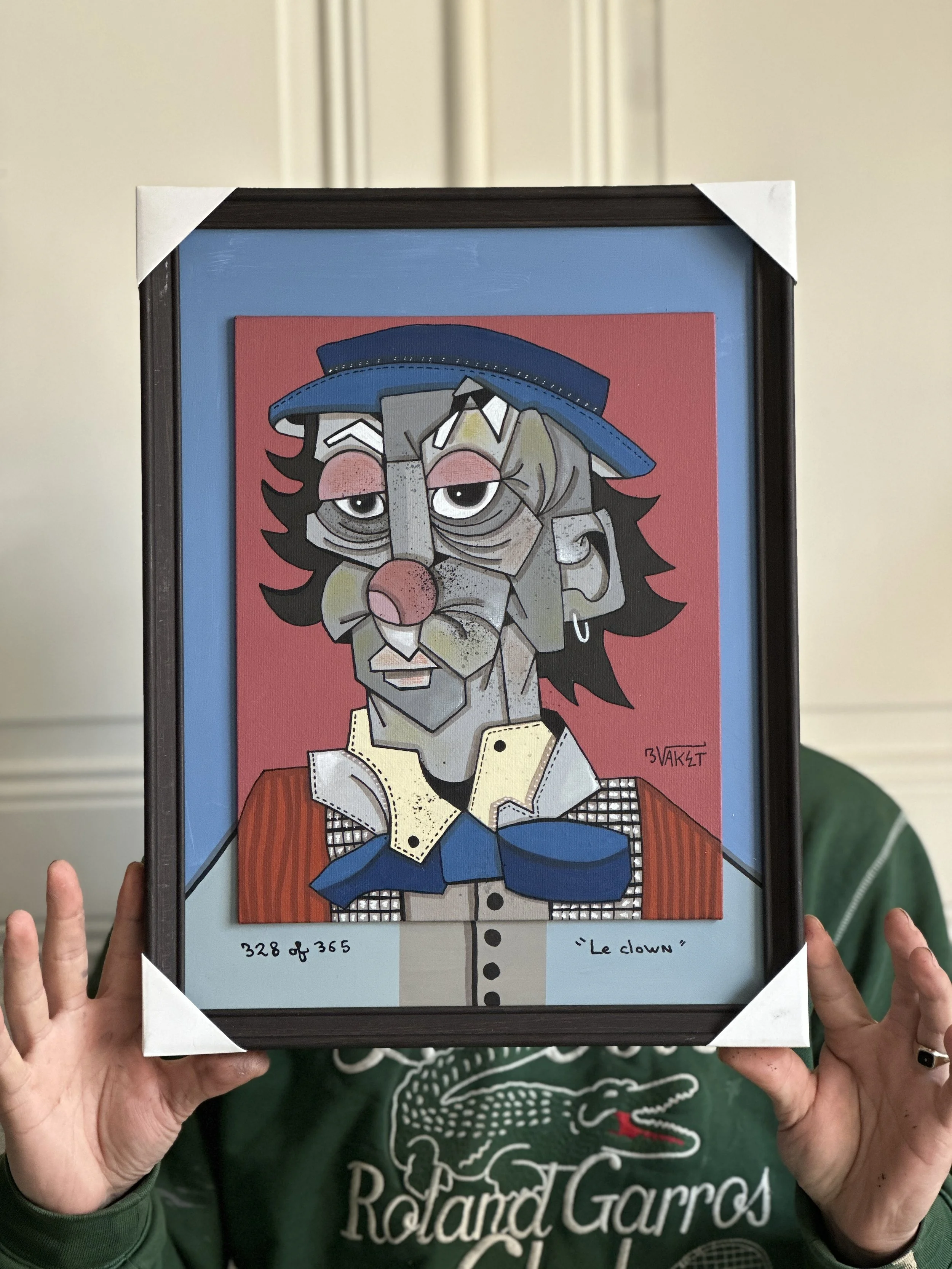 Une personne tient un cadre contenant une illustration de style cubiste d'un clown portant un chapeau bleu, une veste orange, une cravate bleue et une chemise à carreaux. En bas, il y a l'inscription '328 of 365' à gauche et 'Le clowwn' à droite.
