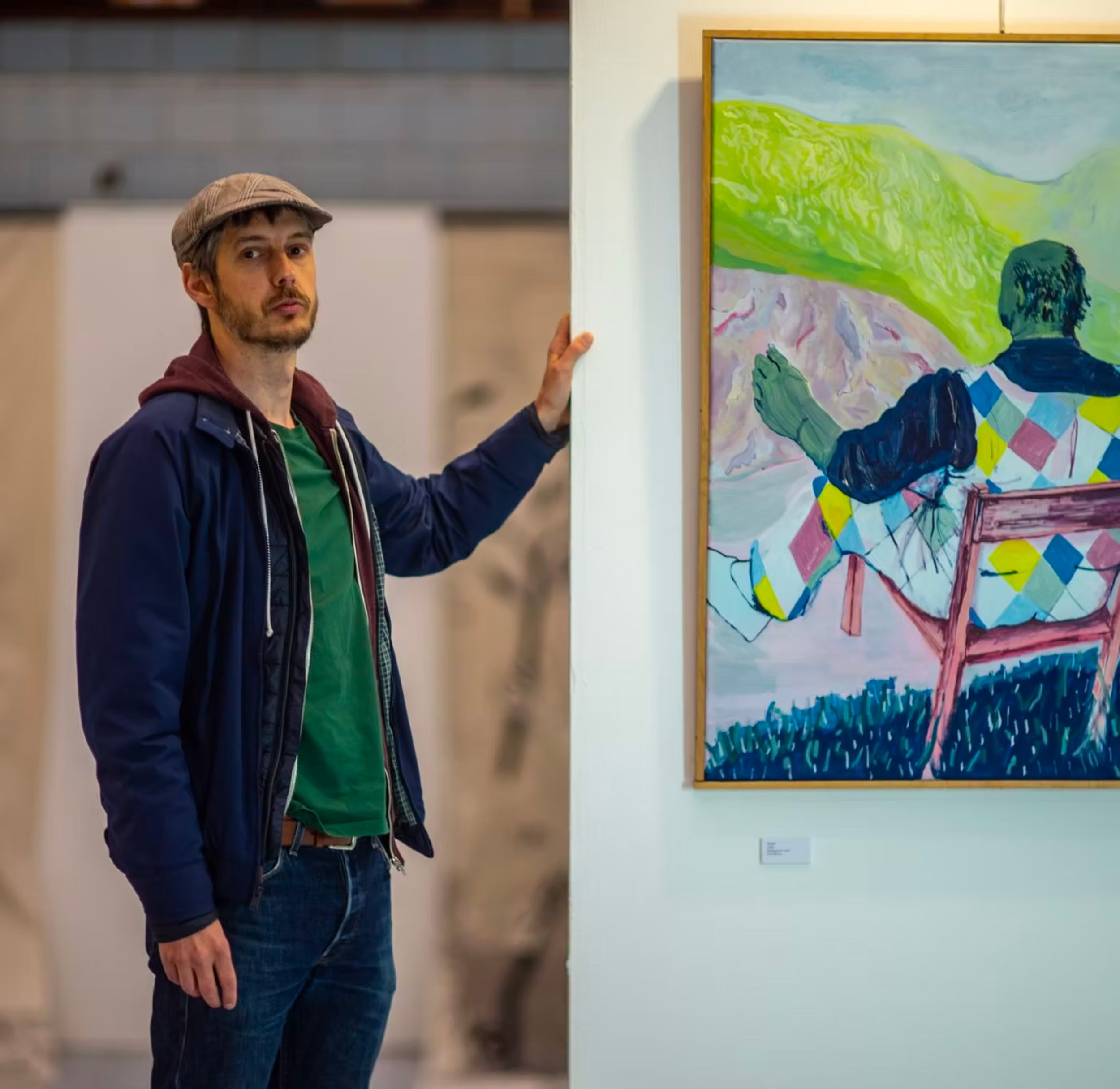 Un homme en veste noire, t-shirt vert et bonnet gris regarde une peinture abstraite dans une galerie
