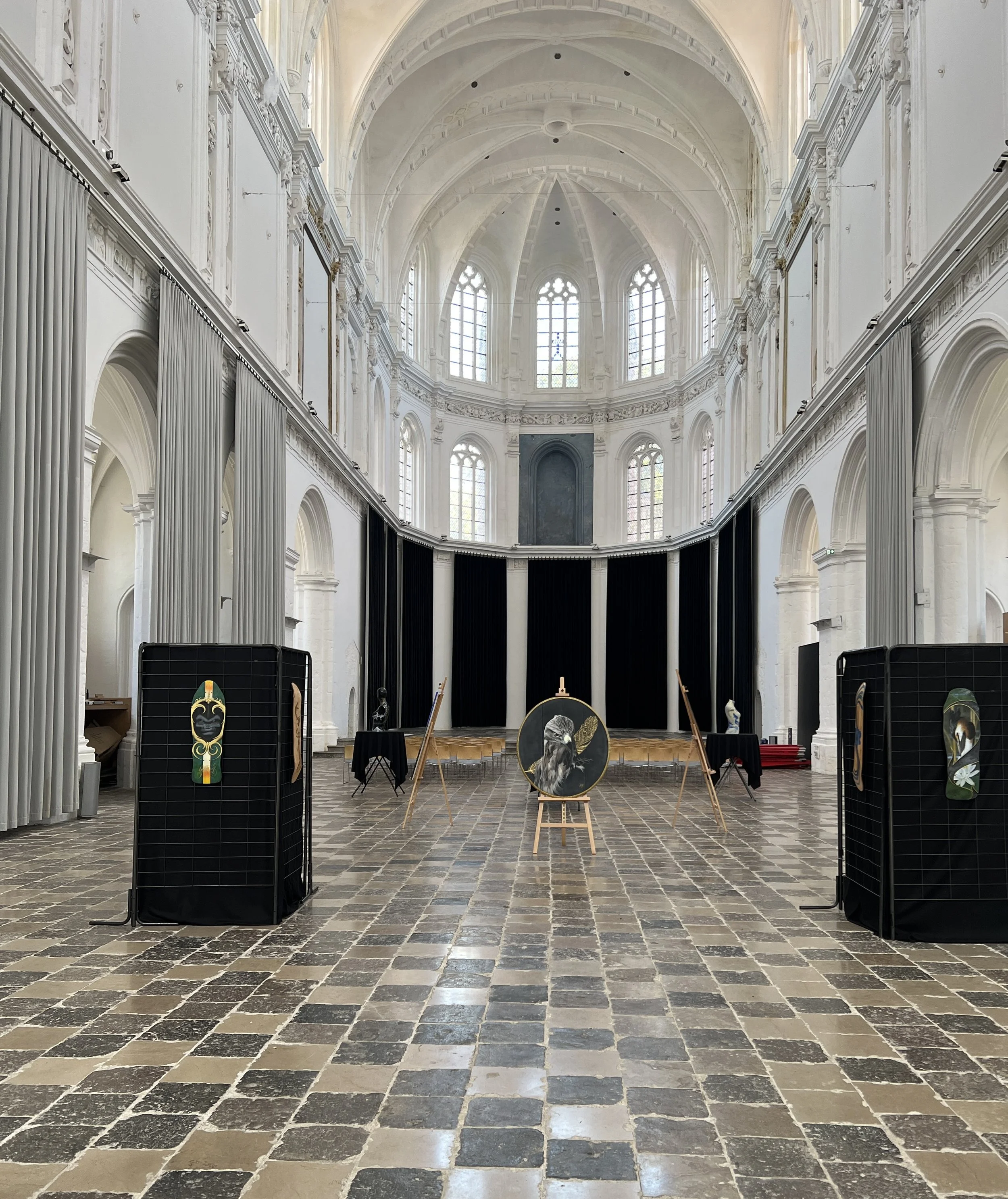Une salle de concert ou d'exposition dans un bâtiment historique avec des vitraux et des décorations classiques, quelques œuvres d'art sont exposées sur des stands et des chevalets au centre.