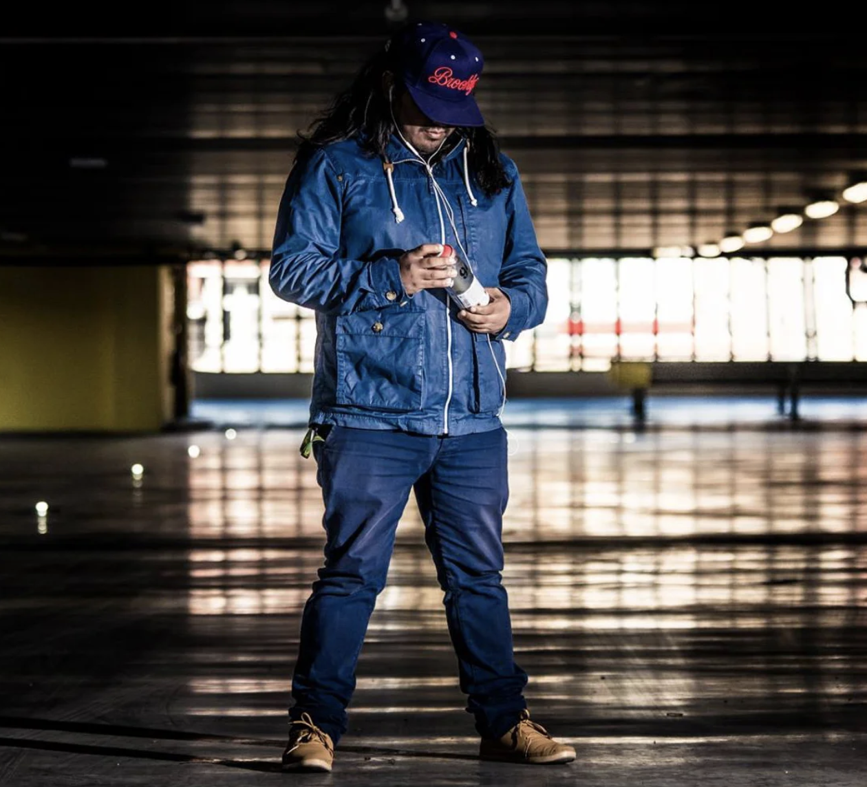 Une personne habillée en bleu, portant une casquette, regarde un objet dans ses mains dans un parking vide avec lumière naturelle de fond.