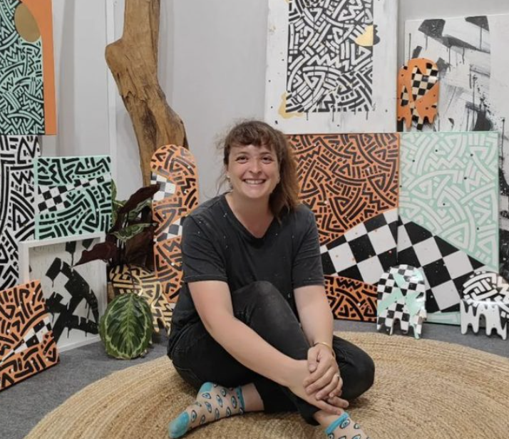 Jeune femme assise sur un tapis en raphia, souriante, entourée d'œuvres d'art colorées en motifs géométriques et abstraits, avec une grande pièce de bois décorative et plusieurs plantes dans un espace d'art.