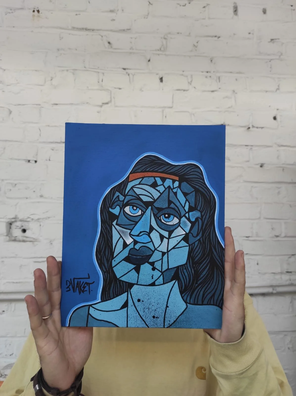 Une personne tient un tableau représentant une œuvre d'art abstraite avec des lignes et des formes géométriques, principalement en nuances de bleu, noir et blanc, avec un fond blanc en pierre.