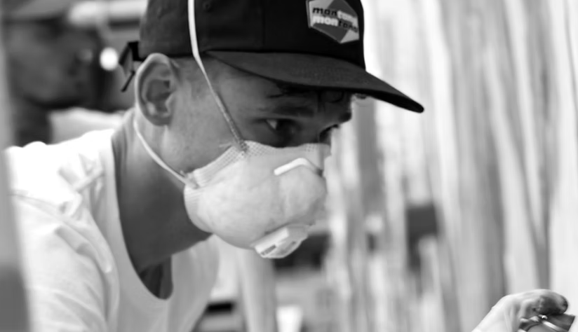 Un homme portant un masque de protection et une casquette regarde attentivement un objet qu'il tient dans sa main. PORTRAIT OF MAZER RESTART