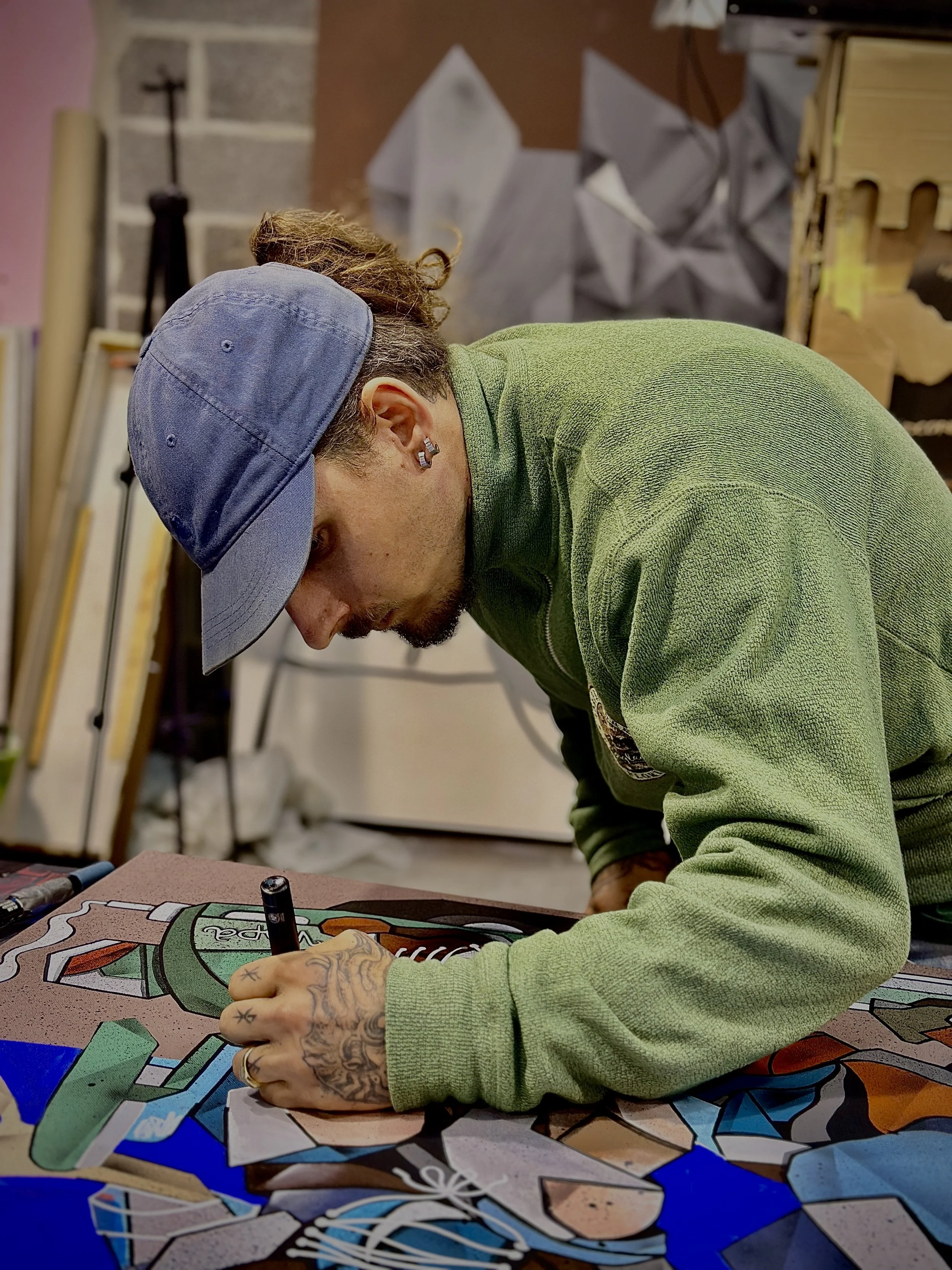 Un homme en casquette noire, portant un sweat vert, dessine une œuvre de street art colorée sur une table, dans un atelier avec des outils et des œuvres en arrière-plan.