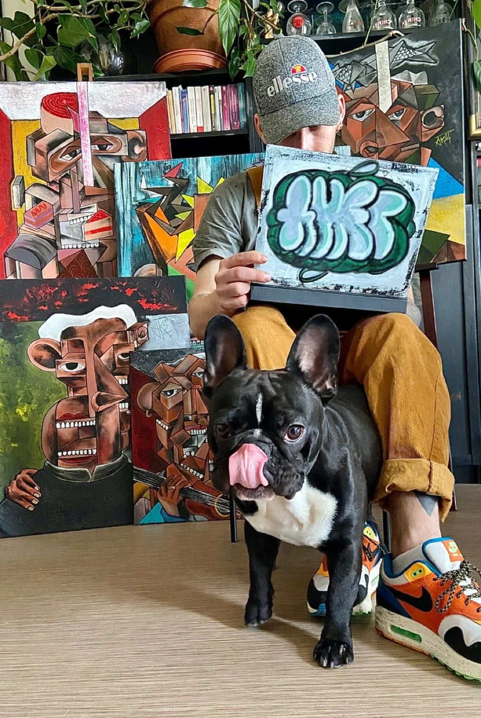 Un homme assis au sol, portant un bonnet gris et des chaussures de sport colorées, tenant une œuvre d'art graffiti, avec des peintures cubistes en arrière-plan et un chien Bouledogue français devant lui, léchant son visage.