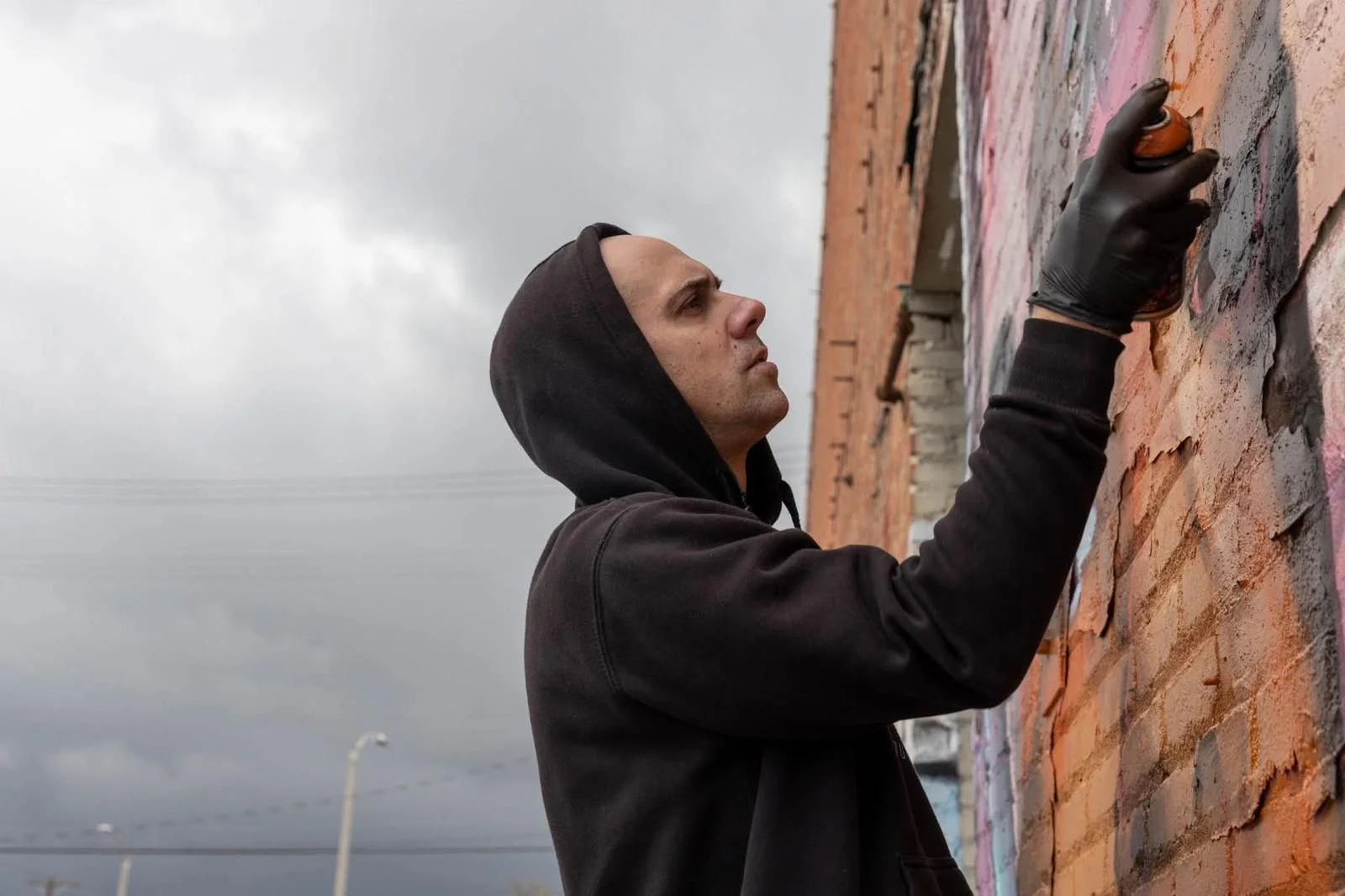 Jeune homme en hoodie noir, portant un gant noir, réalise un graffiti sur un mur en briques. Le ciel est nuageux et il semble concentré dans son activité artistique. PORTRAIT OF DIRTY6