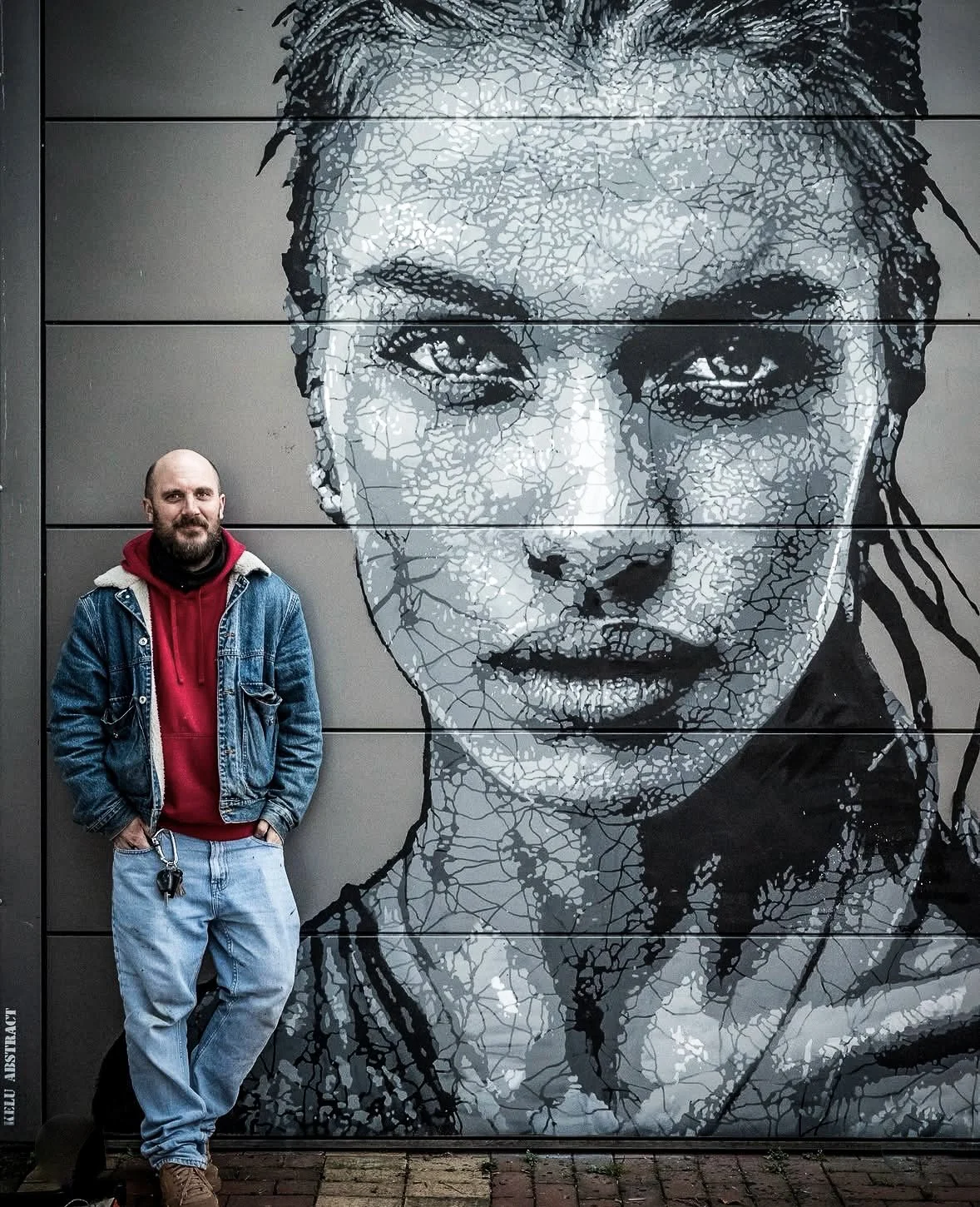 Un homme avec un jean, un sweat à capuche rouge et une veste en jean, se tenant devant une grande fresque murale de visage féminin en noir et blanc. PORTRAIT OF KELU ABSTRACT