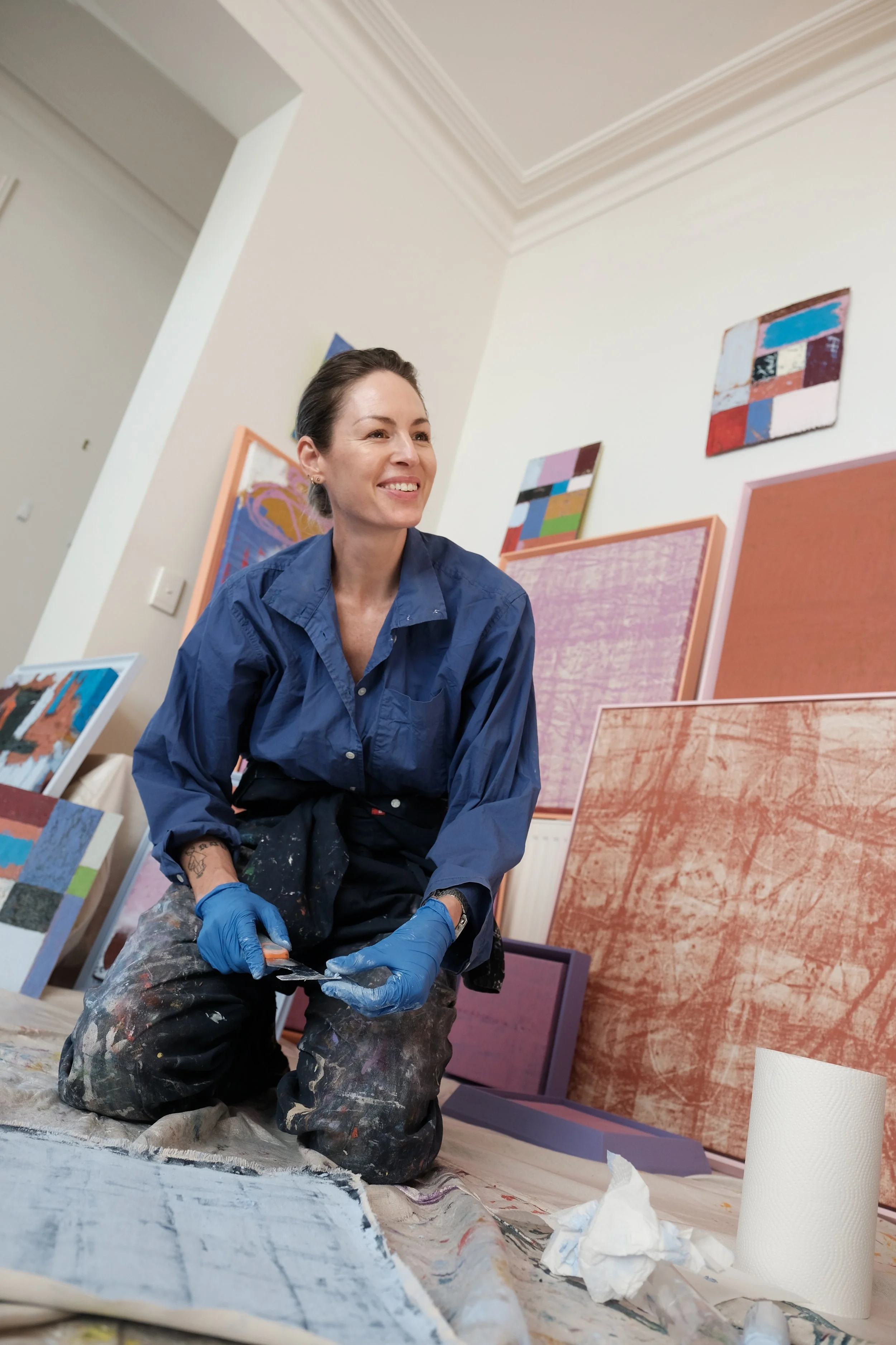 Artist, Toni Vallance, kneeling on the floor, wearing a blue shirt, black paint-splattered pants, and blue gloves, smiling while working on paintings in an art studio.