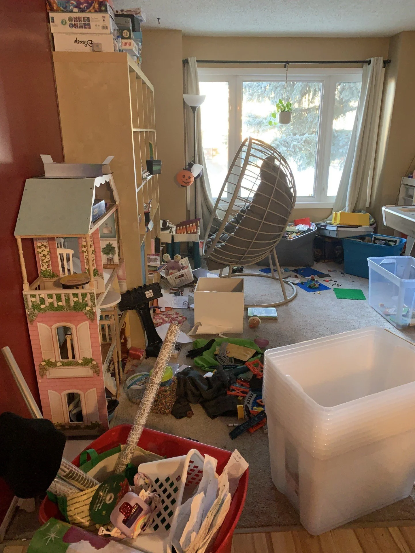 A cluttered indoor room with toys, storage boxes, and furniture scattered across the floor. There is a large window with curtains allowing natural light into the room, a hanging plant, and a white chair near the window. Various boxes and toys are visible, including a pink dollhouse, a large clear plastic storage container, and scattered small objects.