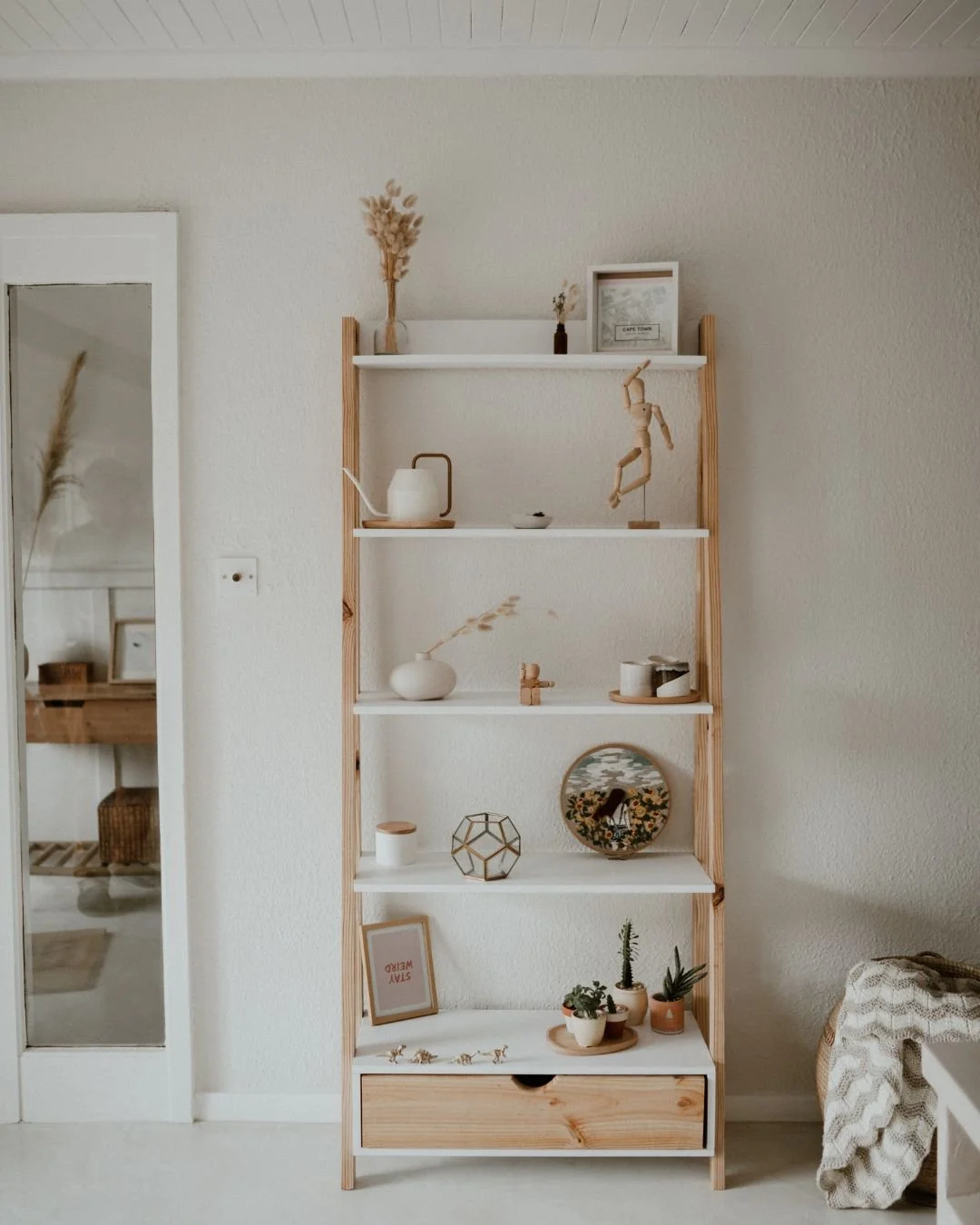A tall wooden and white shelf with decorative items, plants, and framed pictures, set against a white wall in a cozy room.