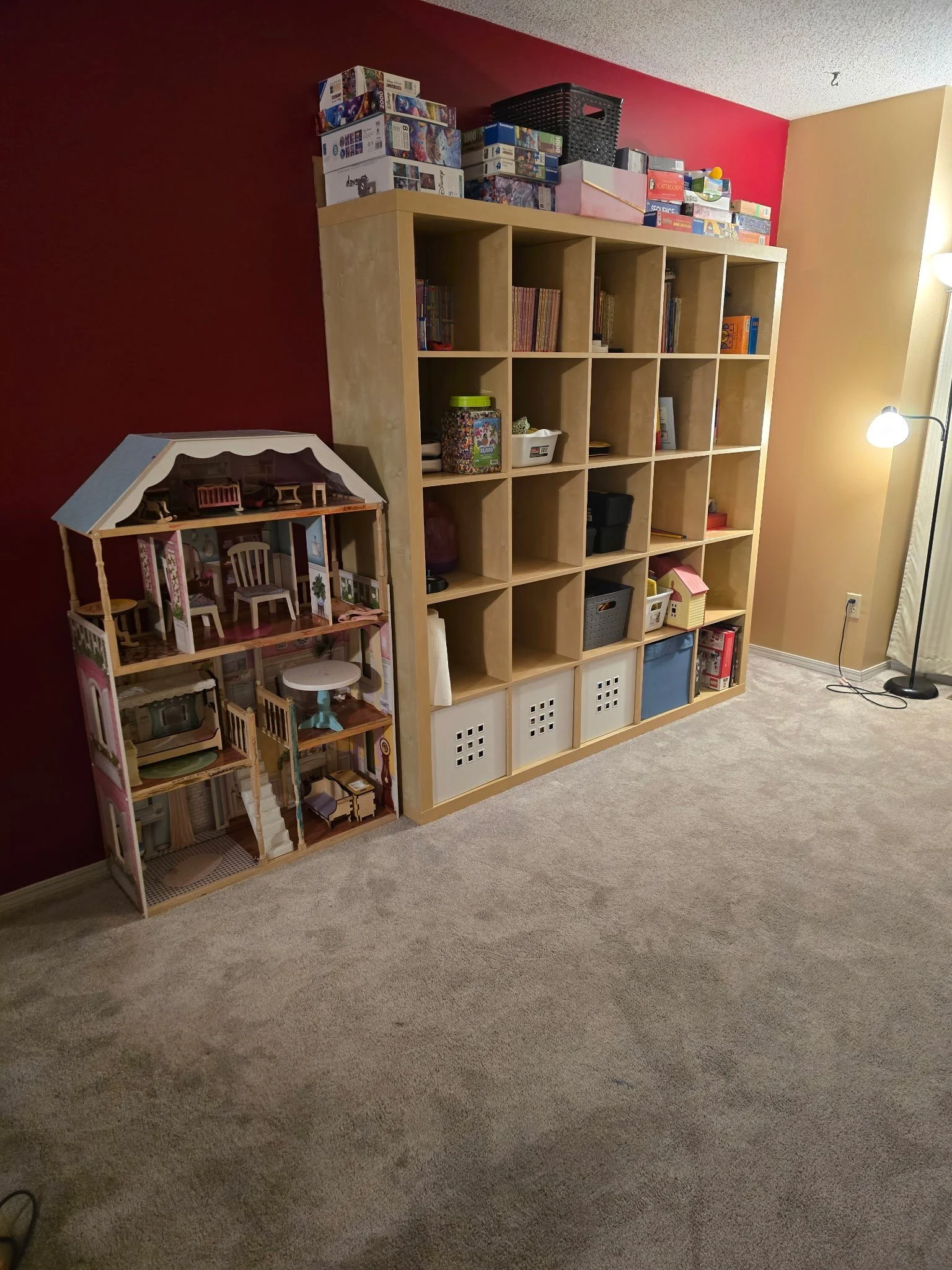 A large wooden bookshelf filled with books and storage bins, with a dollhouse to its left, against a two-tone red and yellow wall, with a floor lamp on the right.