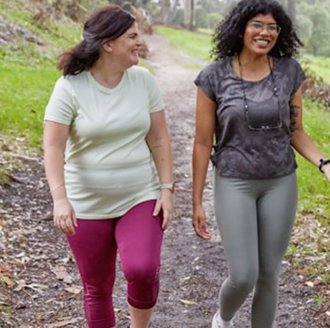 Two women walking outdoors on a dirt trail, smiling and talking, surrounded by greenery.