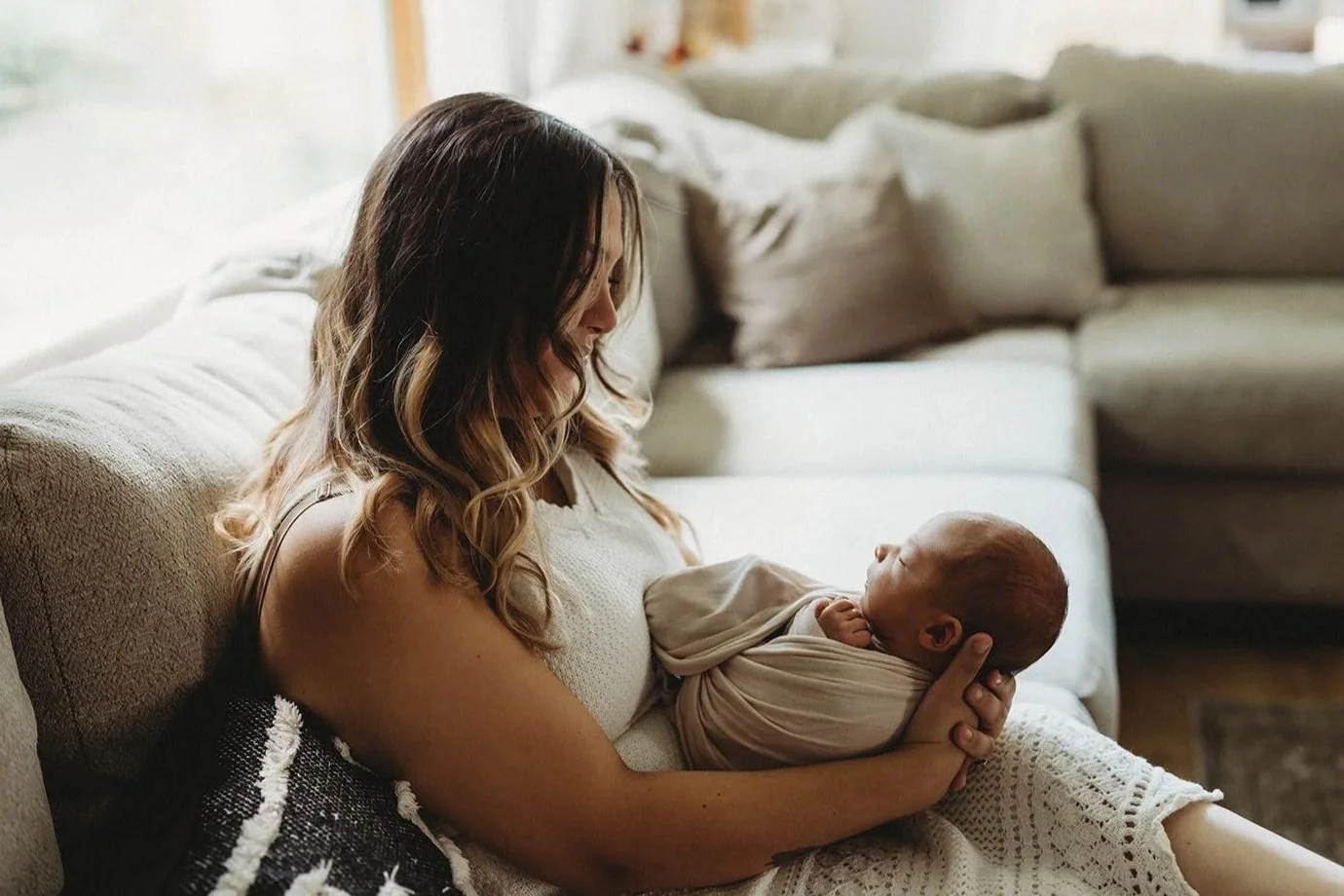A woman with long, wavy hair holding a newborn baby wrapped in a beige blanket on a beige sofa in a cozy living room.