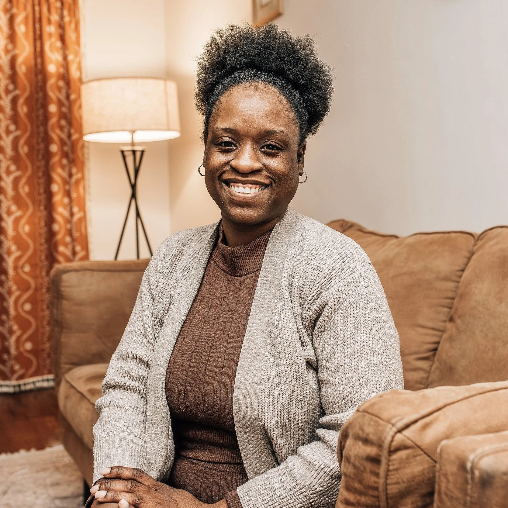 A woman sitting on a beige couch in a living room, smiling at the camera, wearing a brown top and a gray cardigan. There is a lamp and curtain in the background.
