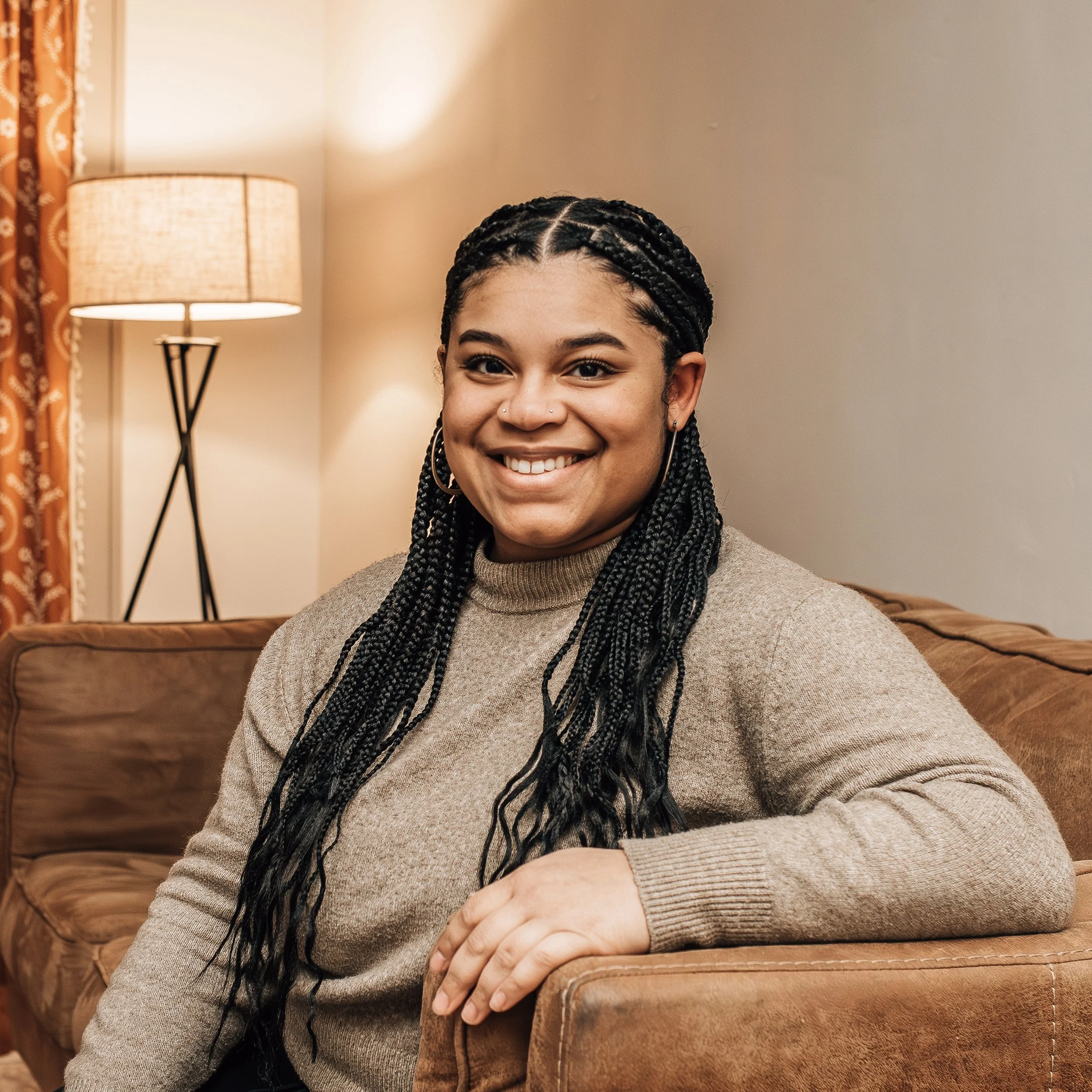 A young woman with braided hair sitting on a brown couch in a cozy living room, smiling at the camera. The room has a lamp and patterned curtains in the background.
