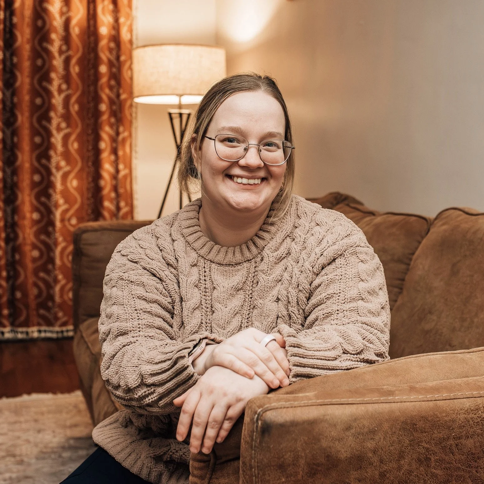 A young woman with glasses and long hair smiling while sitting on a brown sofa in a cozy living room, with a lamp and curtains in the background.