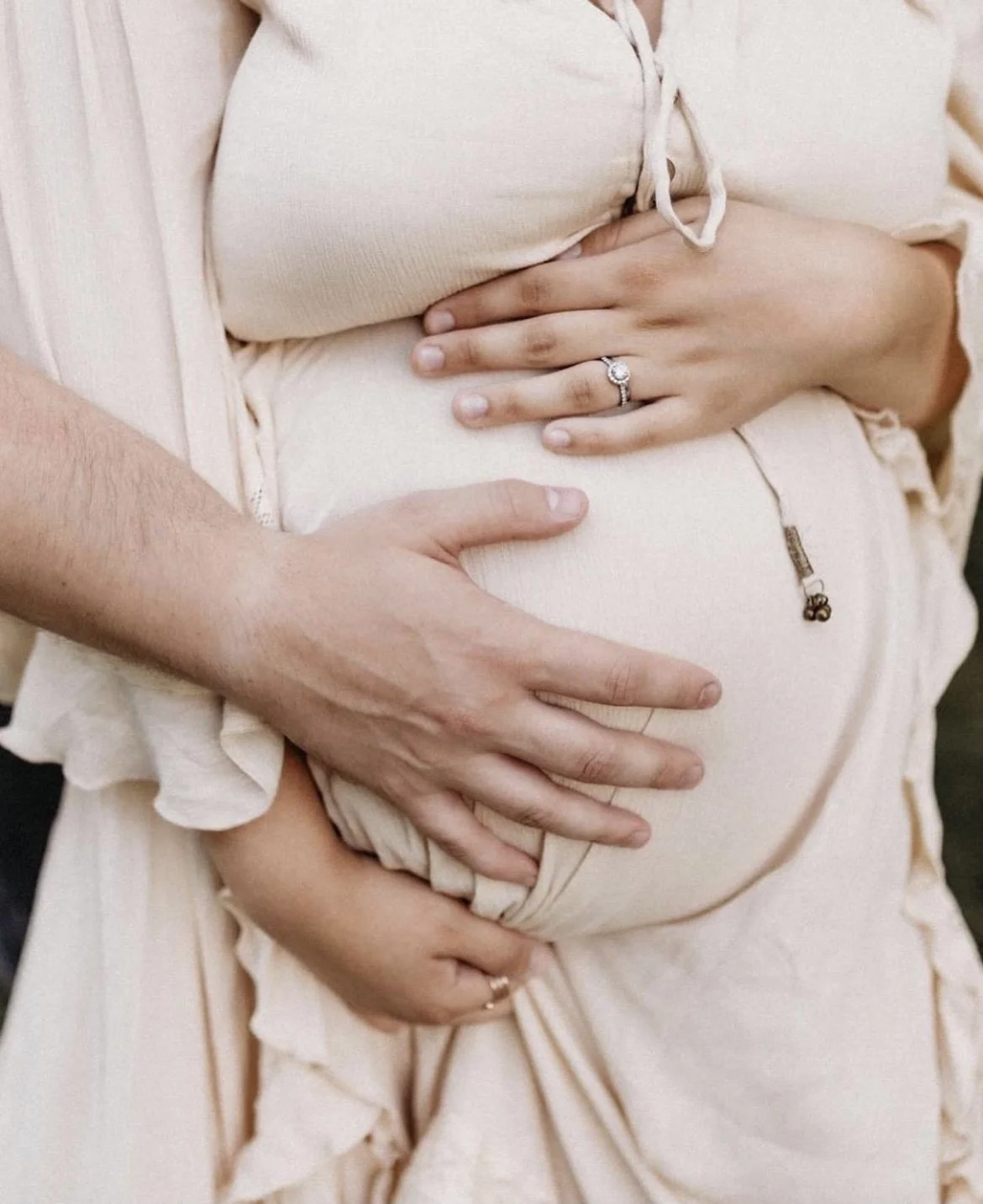 Close-up of a pregnant woman's belly with two hands, one of a man and one of a woman, gently caressing it. The woman's hand features an engagement ring. The woman is wearing a cream-colored outfit with a drawstring.