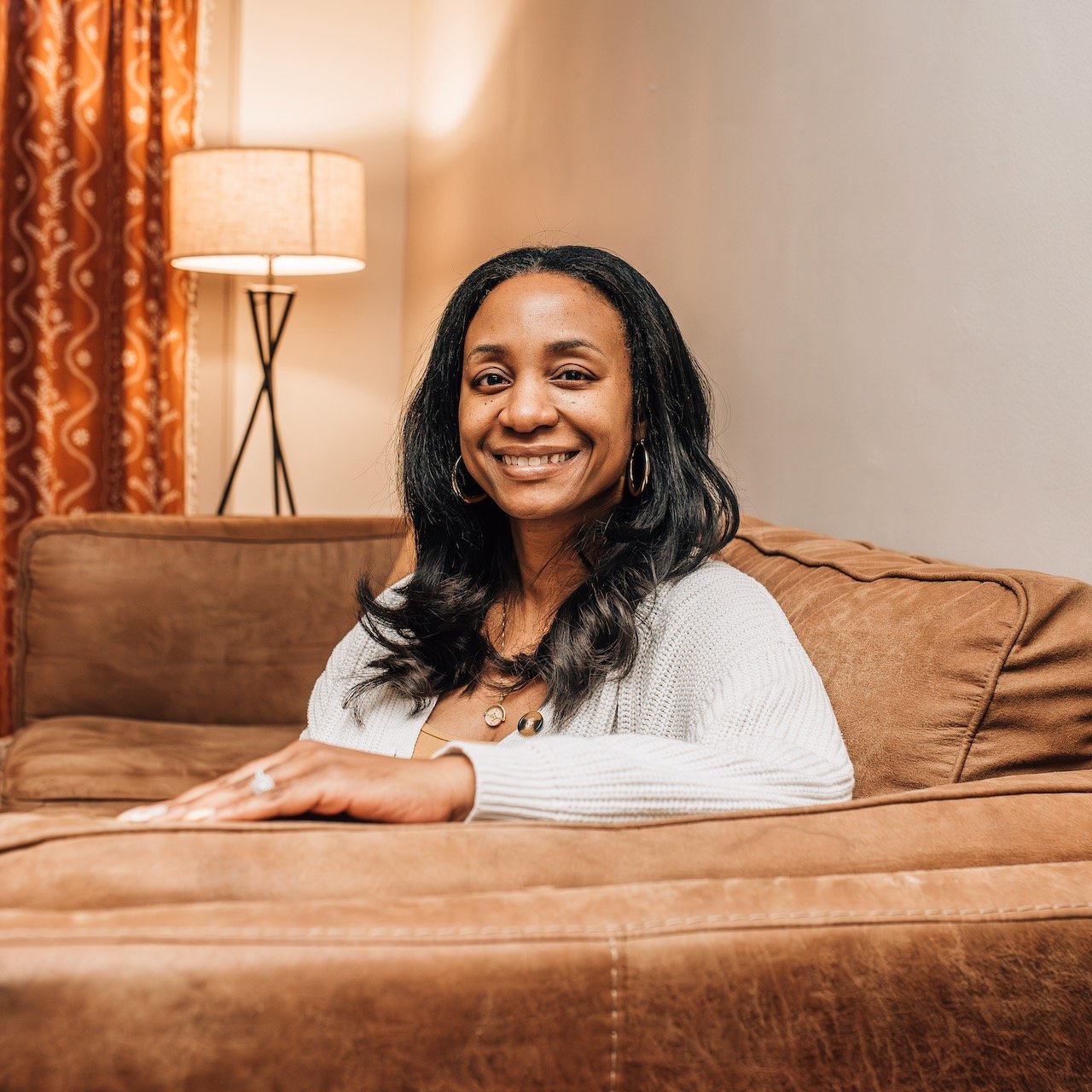 A woman with long dark hair smiling, sitting on a brown couch in a warmly lit room with beige walls, a lamp, and patterned curtains in the background.