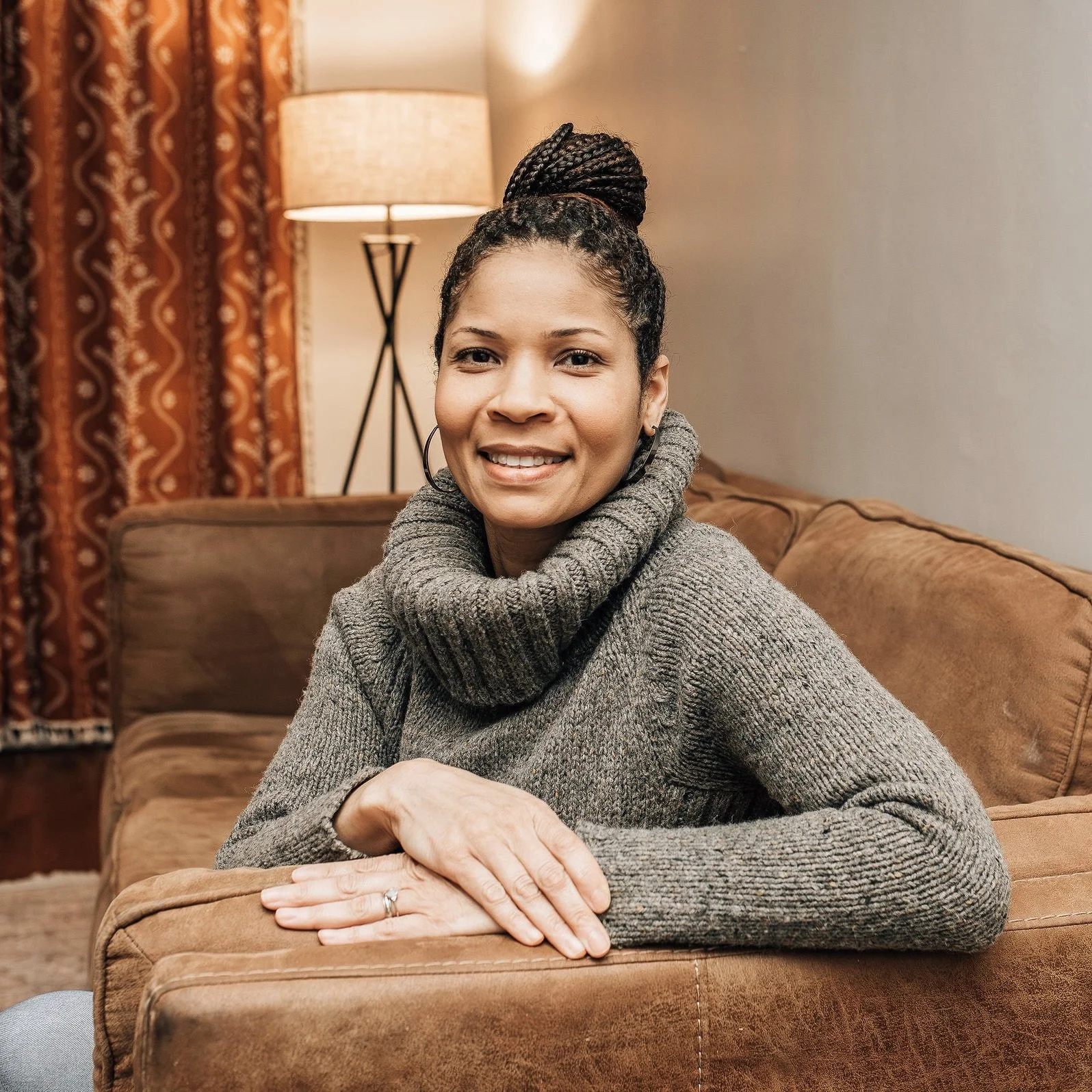 A woman with braids in a bun, wearing a gray turtleneck sweater, smiling while sitting on a brown couch in a cozy living room with a lamp and patterned curtains in the background.