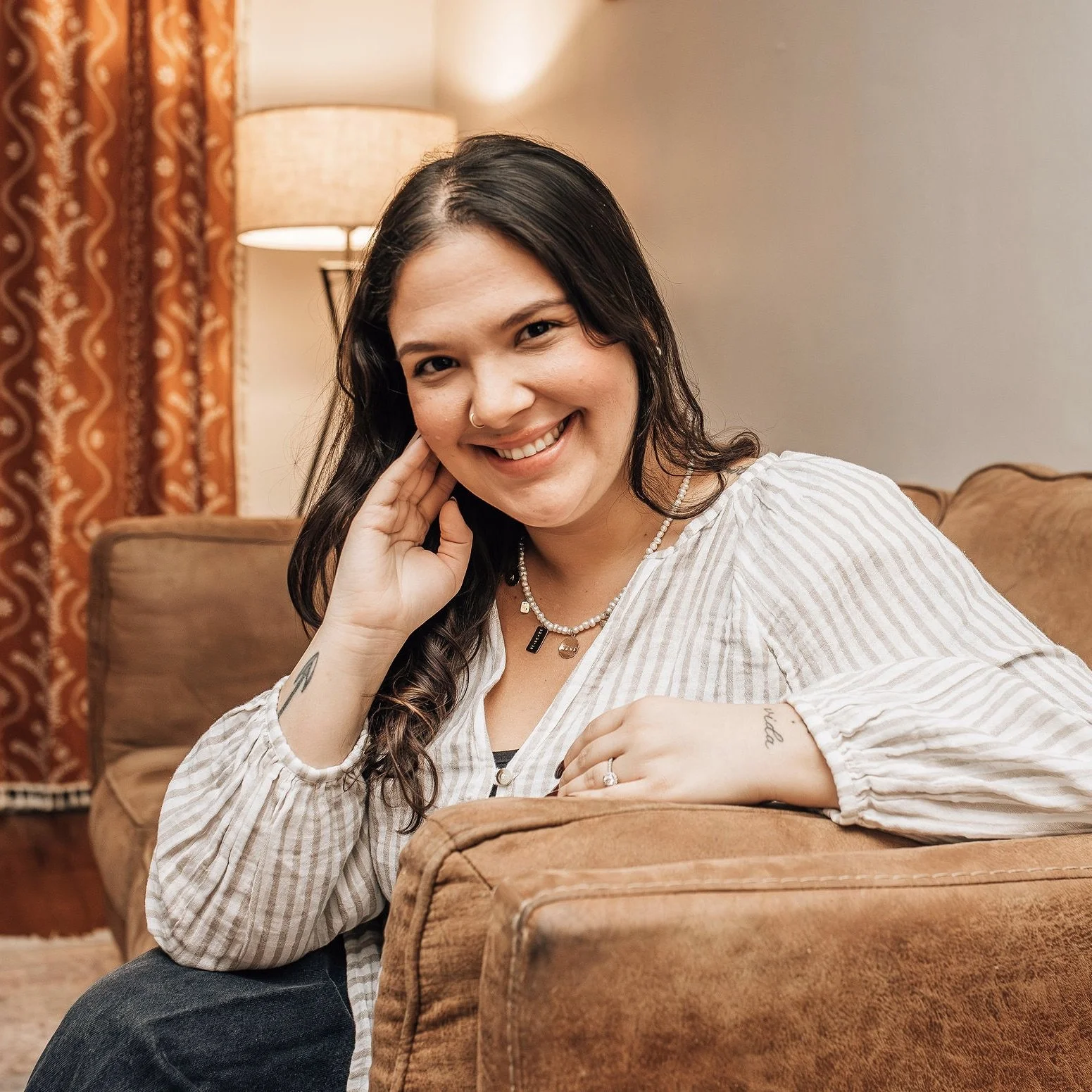 A woman with dark hair, wearing a striped white and beige shirt and jewelry, sitting on a brown couch in a cozy living room, smiling at the camera.