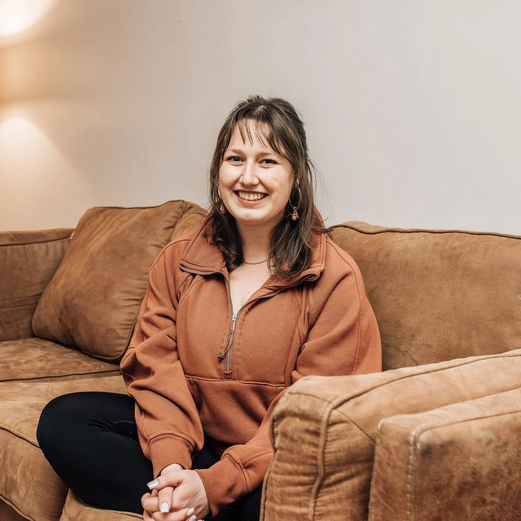 A young woman with shoulder-length brown hair and light skin, smiling while sitting on a tan couch with matching pillows, wearing a rust-colored zip-up jacket and black pants, in a cozy indoor setting.