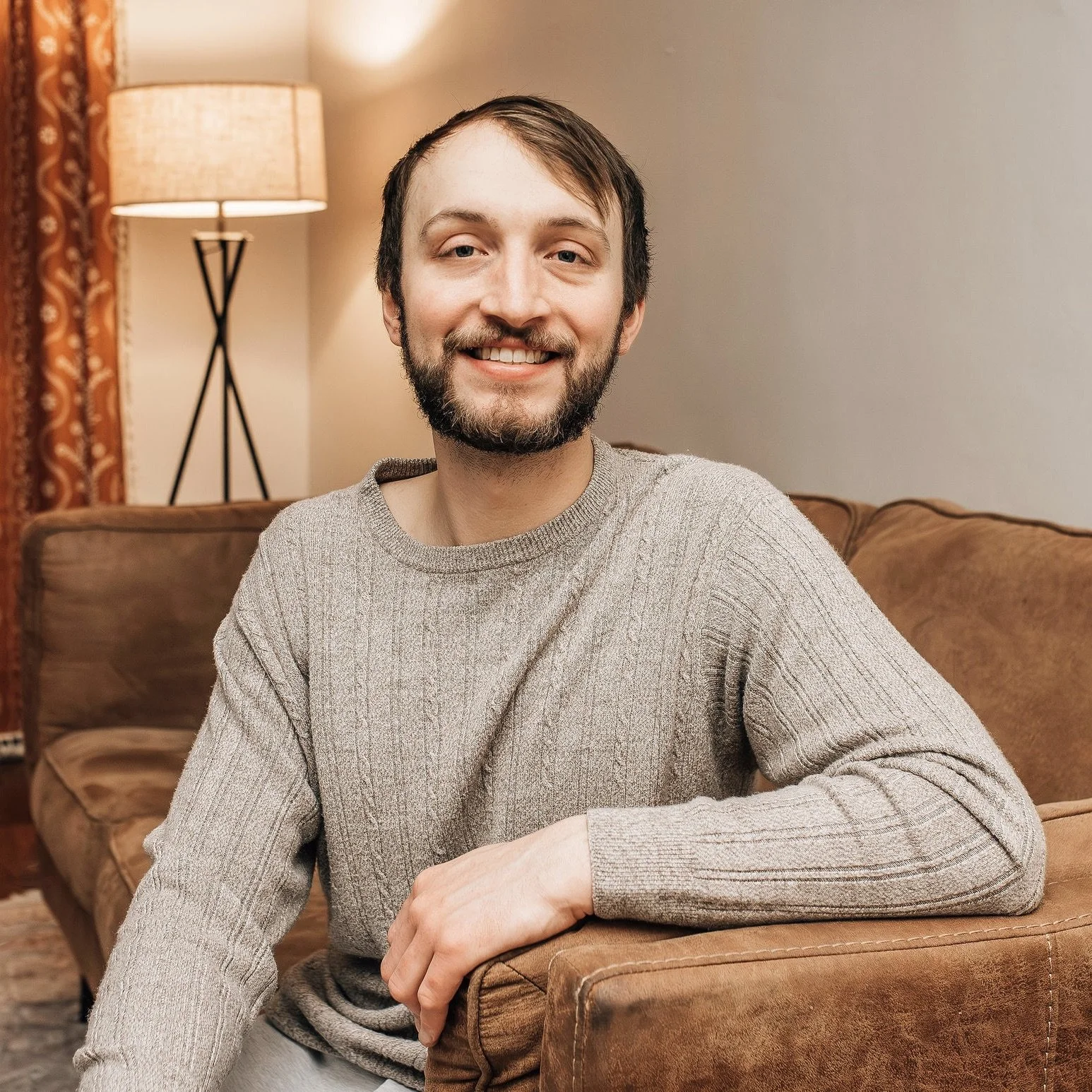 A smiling man with a beard sitting on a brown sofa in a cozy living room, next to a lamp and curtains.