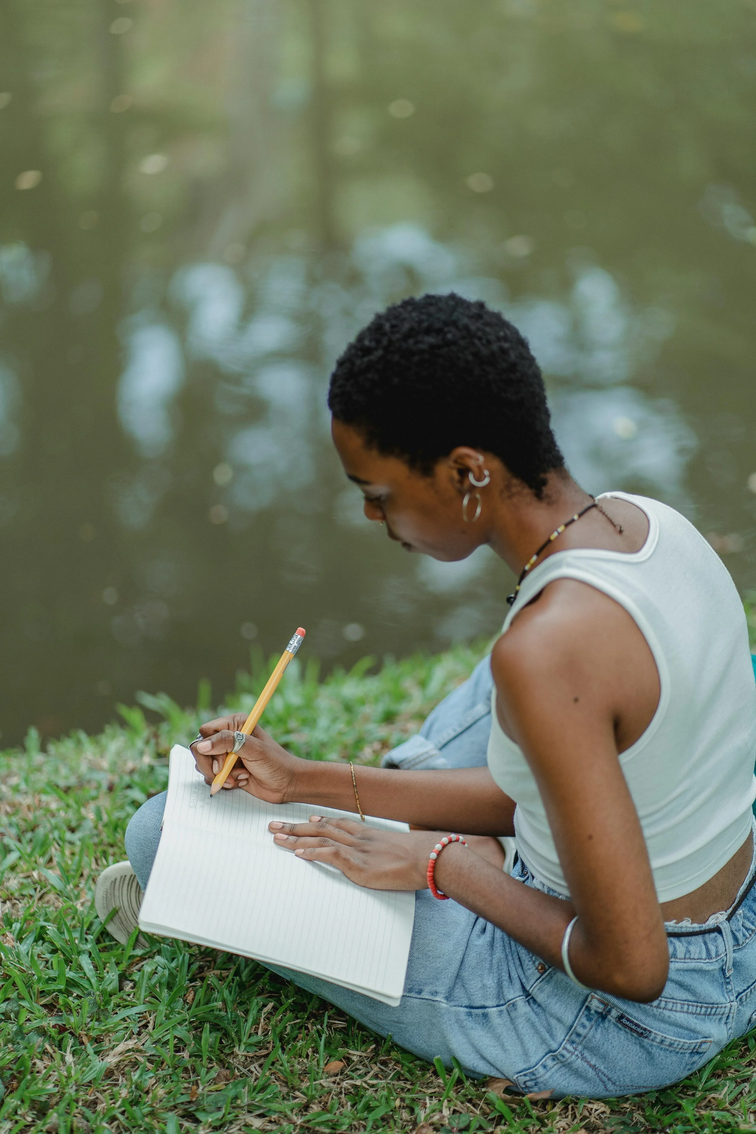 A young woman with short curly hair, wearing a white sleeveless top and jeans, sitting on grass near a pond, writing in a notebook with a pencil.
