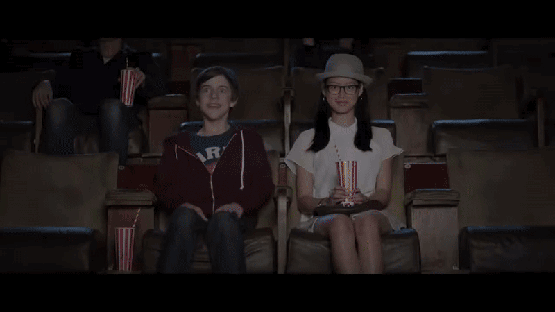 Two children sitting in a movie theater, watching a film, with popcorn and drinks in front of them.