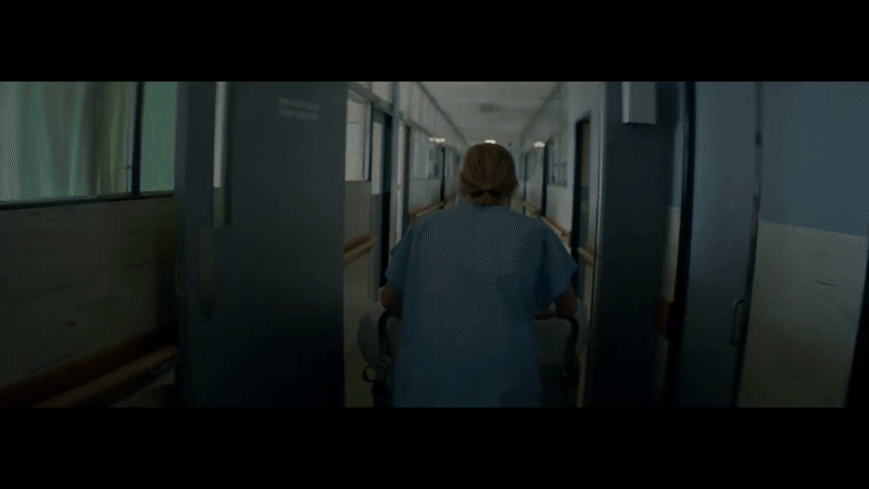 A healthcare worker wearing blue scrubs pushes a mobile medical cart down a hospital corridor.