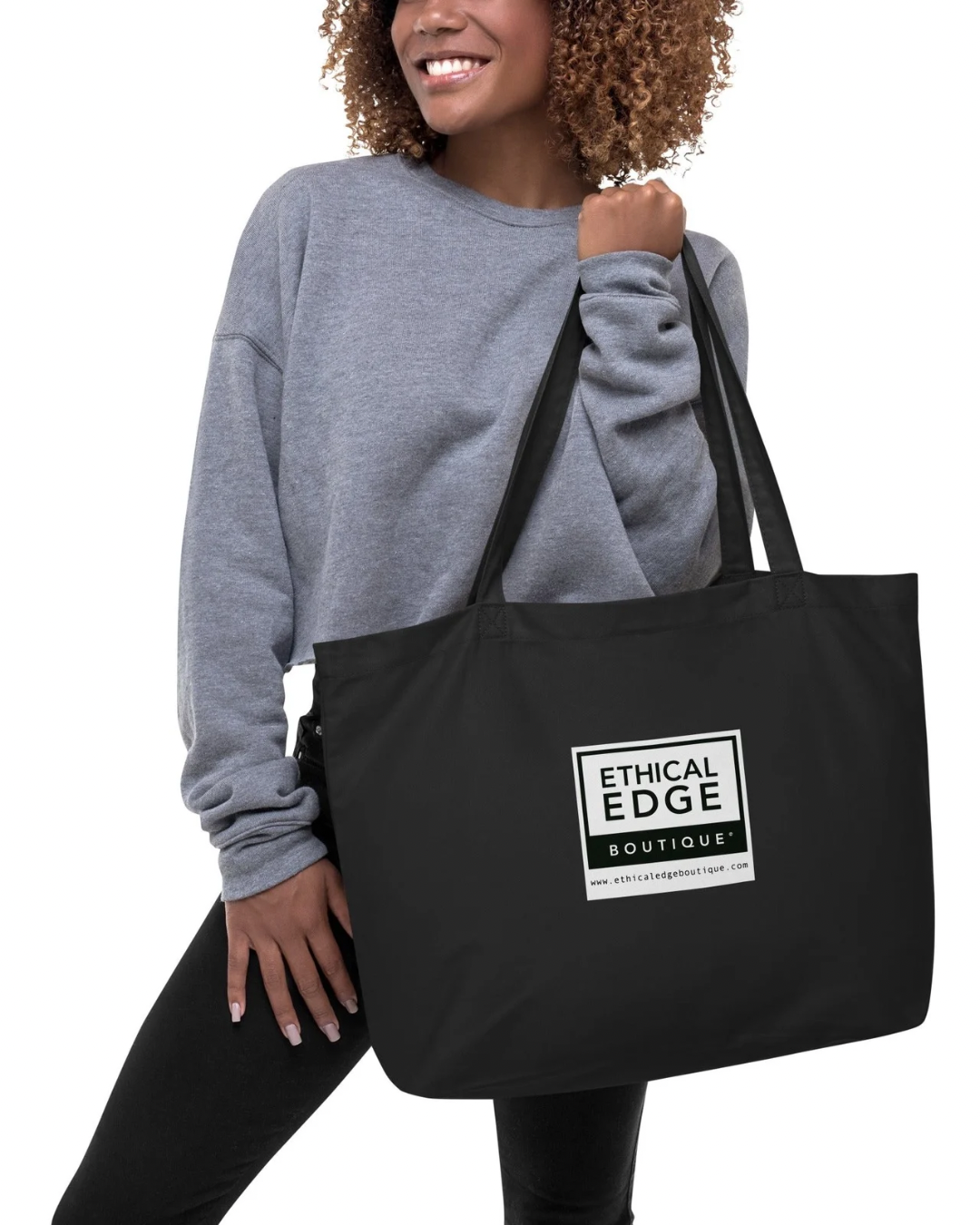 Woman in a gray sweatshirt holding a black tote bag with a logo reading 'Ethical Edge Boutique' on a white background.
