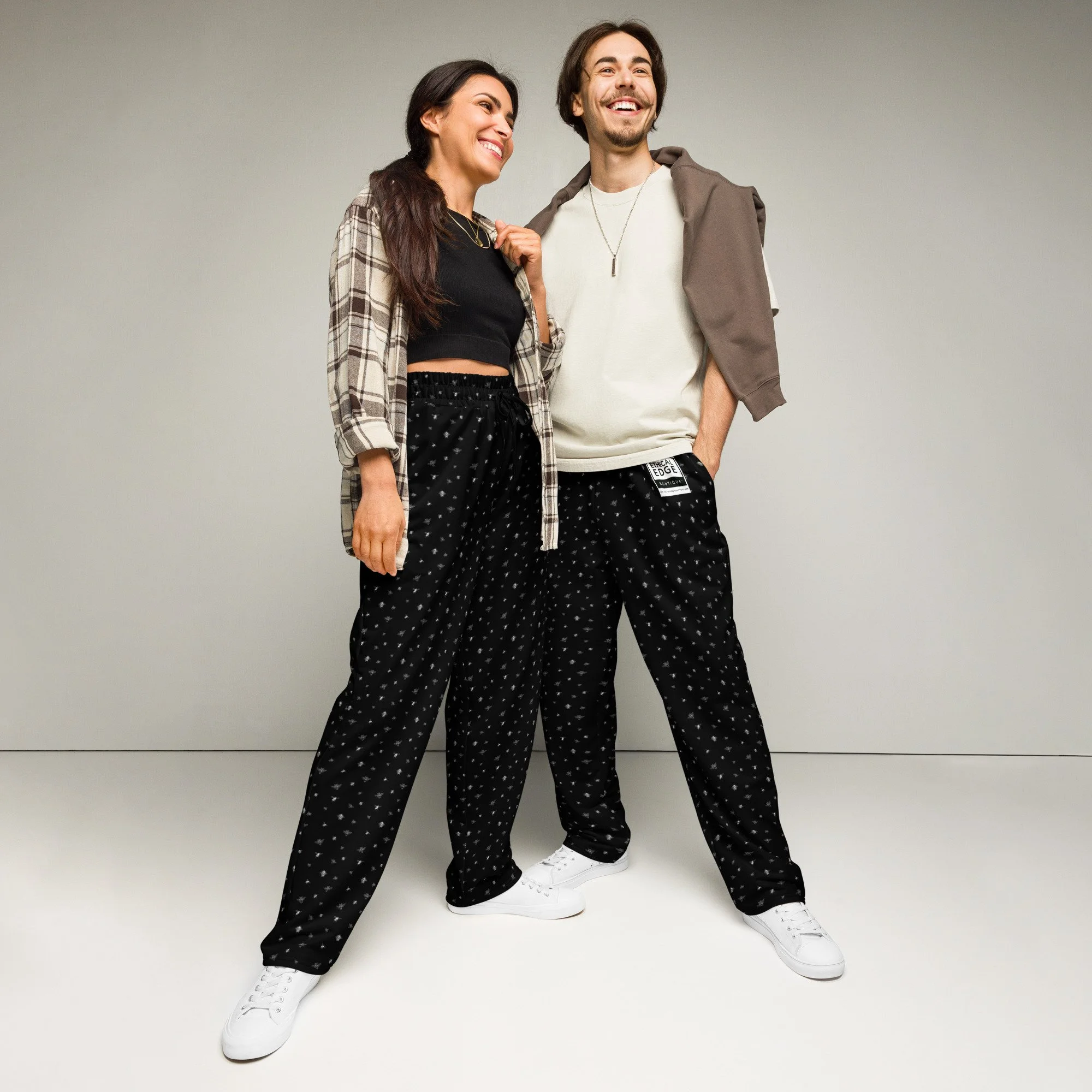 A woman and a man smiling and standing together against a plain background. She is wearing a black crop top, plaid shirt, black patterned pants, and white sneakers. He is wearing a white shirt, black patterned pants, a gray sweatshirt draped over his