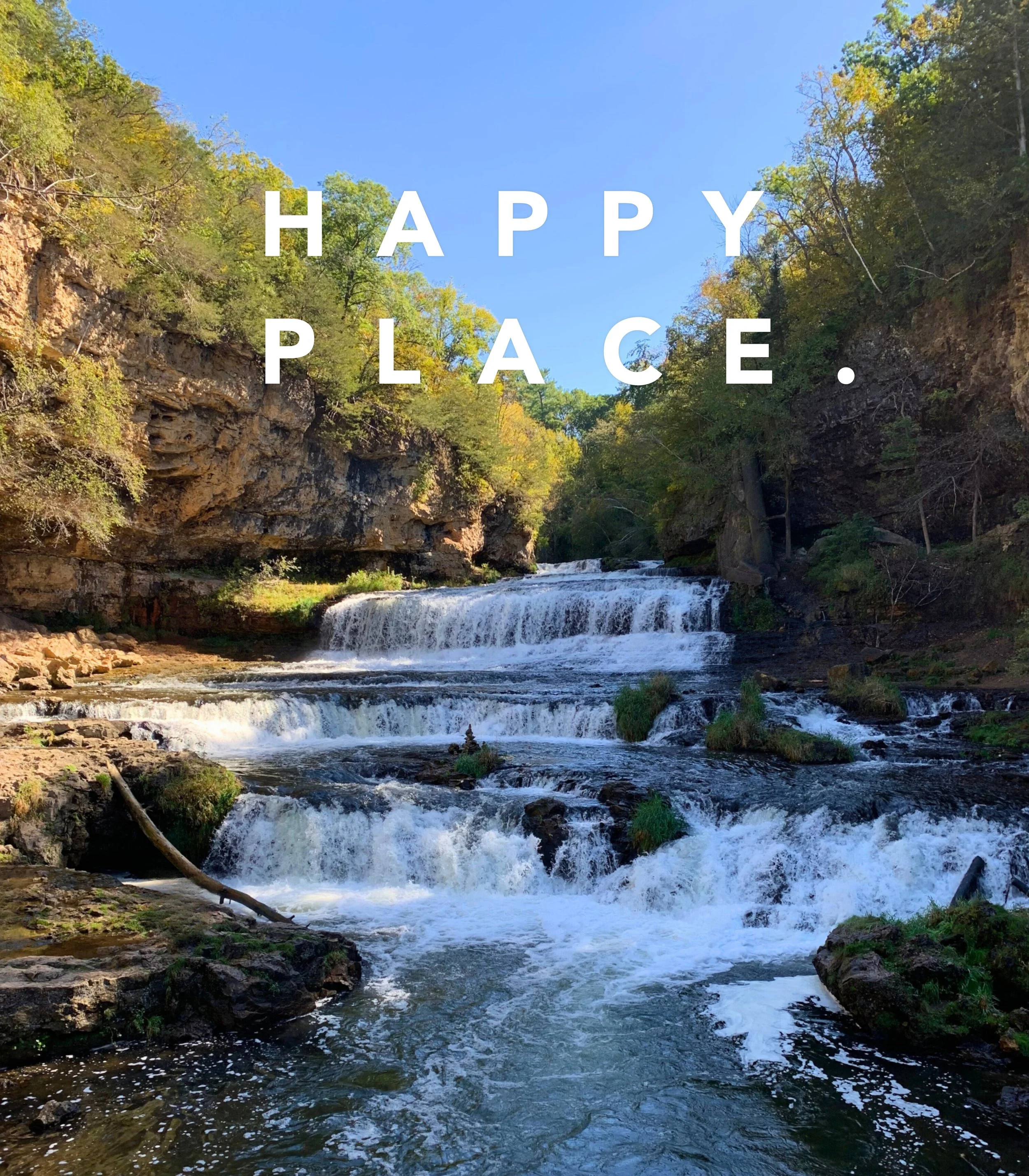 A scenic waterfall flowing through a rocky riverbed surrounded by green trees under a blue sky with the words 'HAPPY PLACE.' in white font