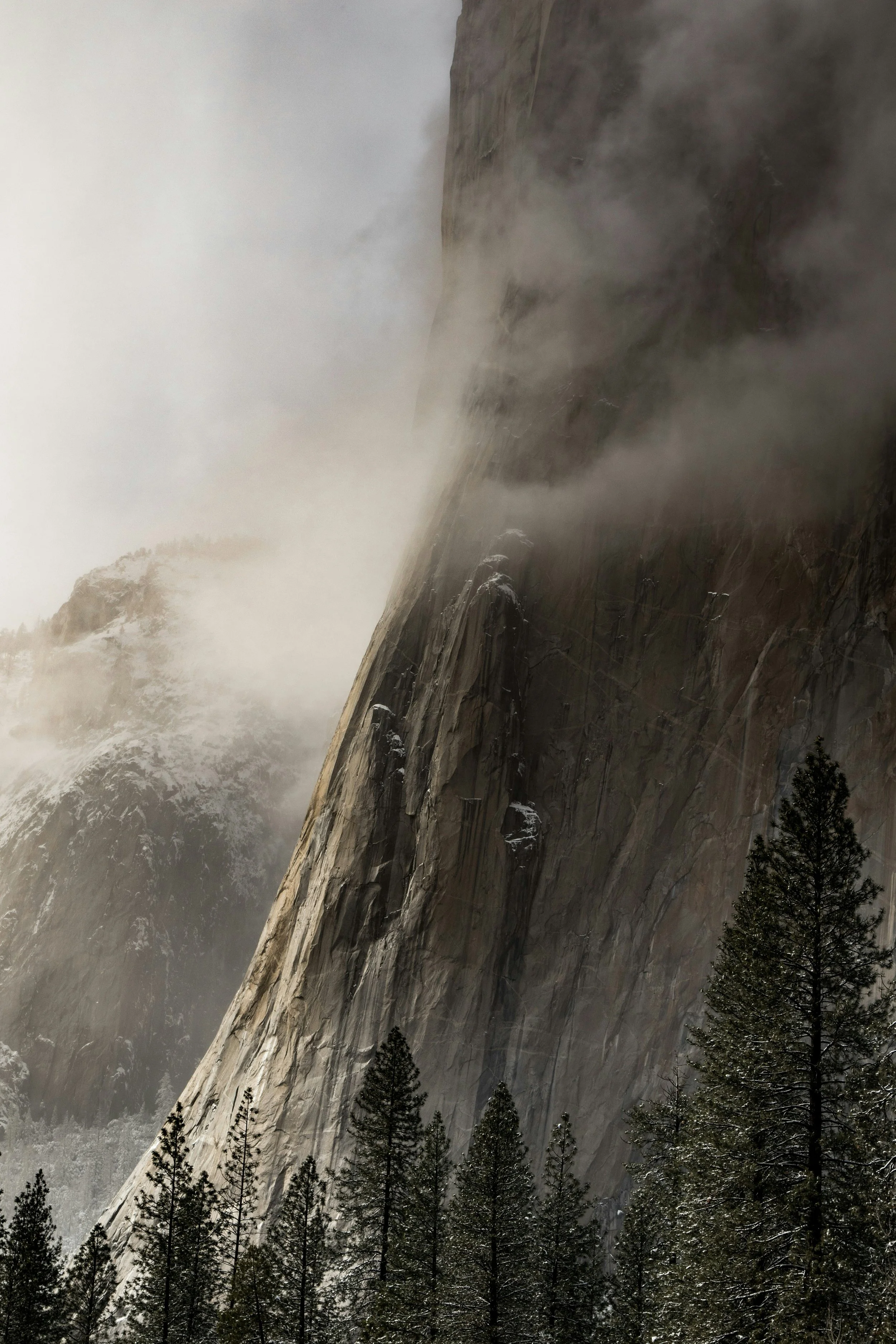 A large mountain with a steep, rocky slope partially covered by mist and clouds, with trees at the base.