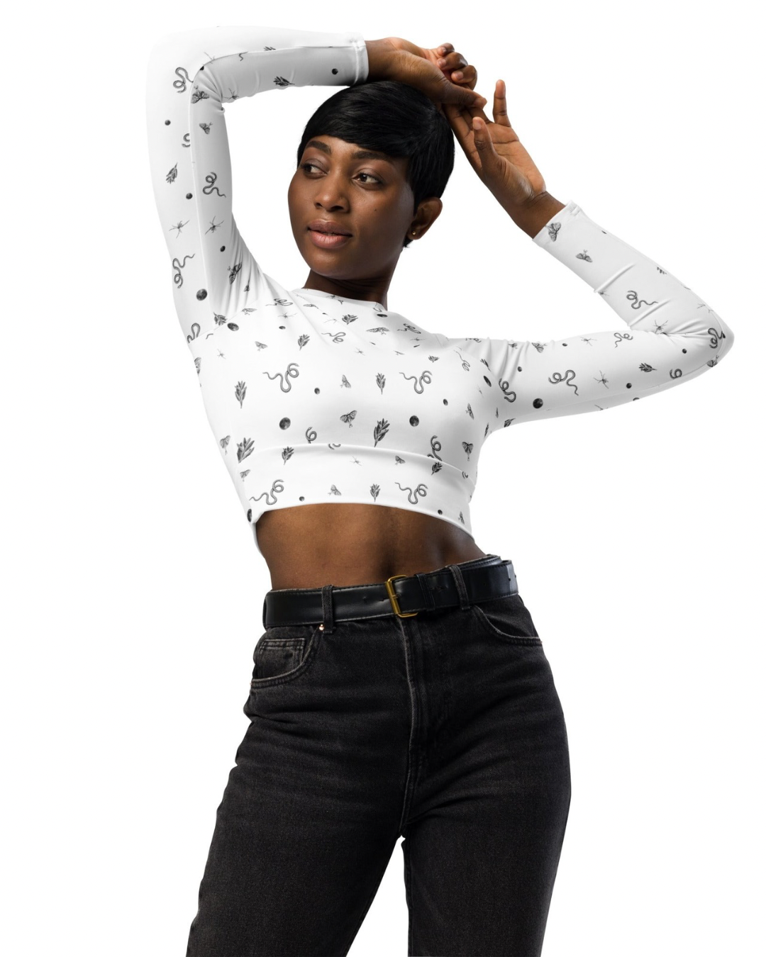 A woman with short black hair wearing a white, long-sleeve crop top with a black snake and leaf pattern and high-waisted black jeans, posing with arms raised and hands touching her head against a plain white background.