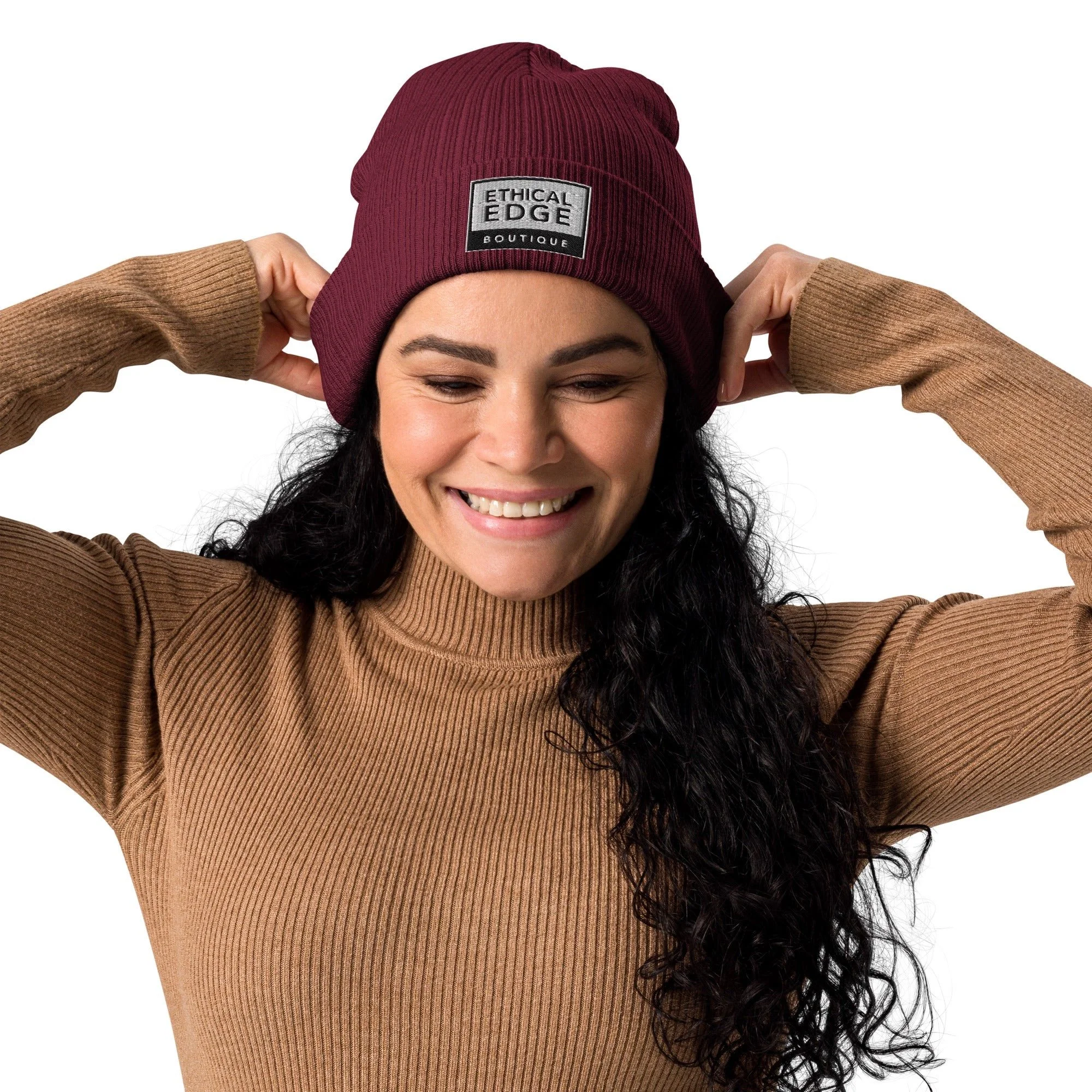 A woman with long curly dark hair smiling while wearing a maroon beanie and a brown ribbed turtleneck sweater.