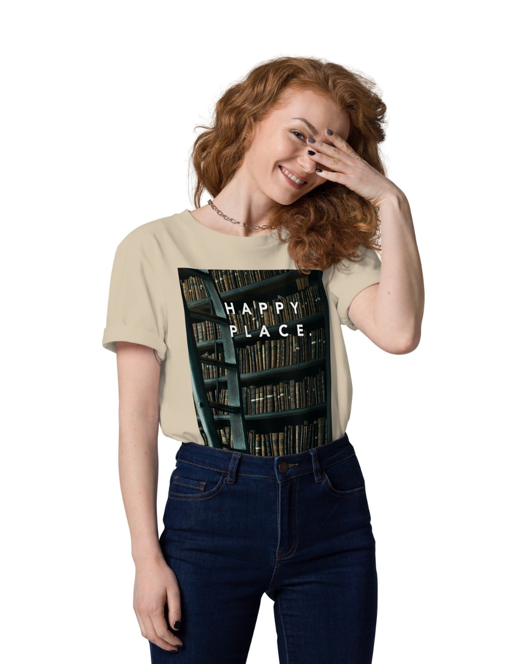 A woman with red curly hair in a beige T-shirt with a bookshelf print and the words 'HAPPY PLACE,' smiling and covering one eye with her hand.