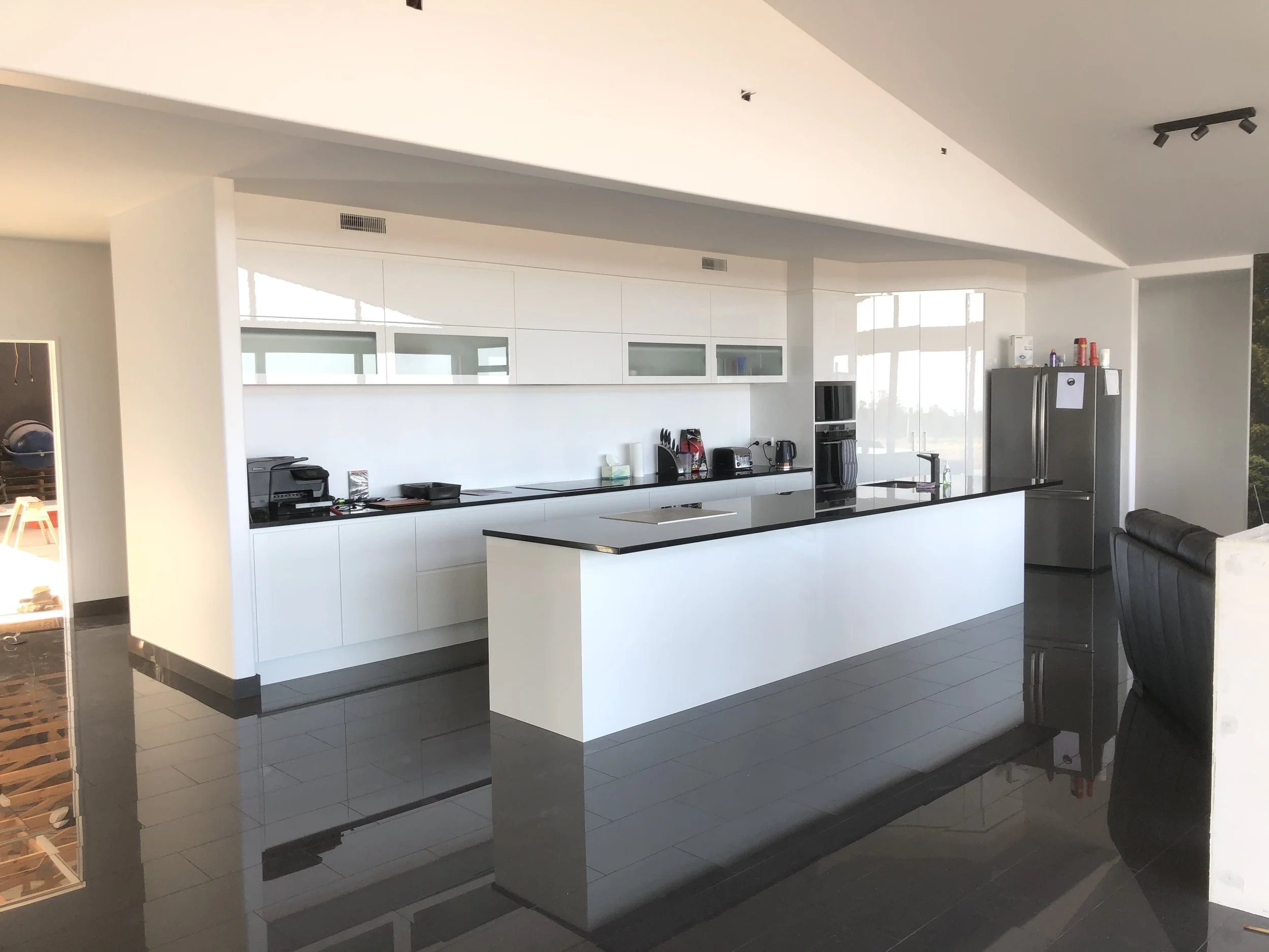 Modern kitchen with white cabinets, black countertops, and stainless steel appliances, including a refrigerator and oven, with a black tiled floor and a dining area with black chairs.