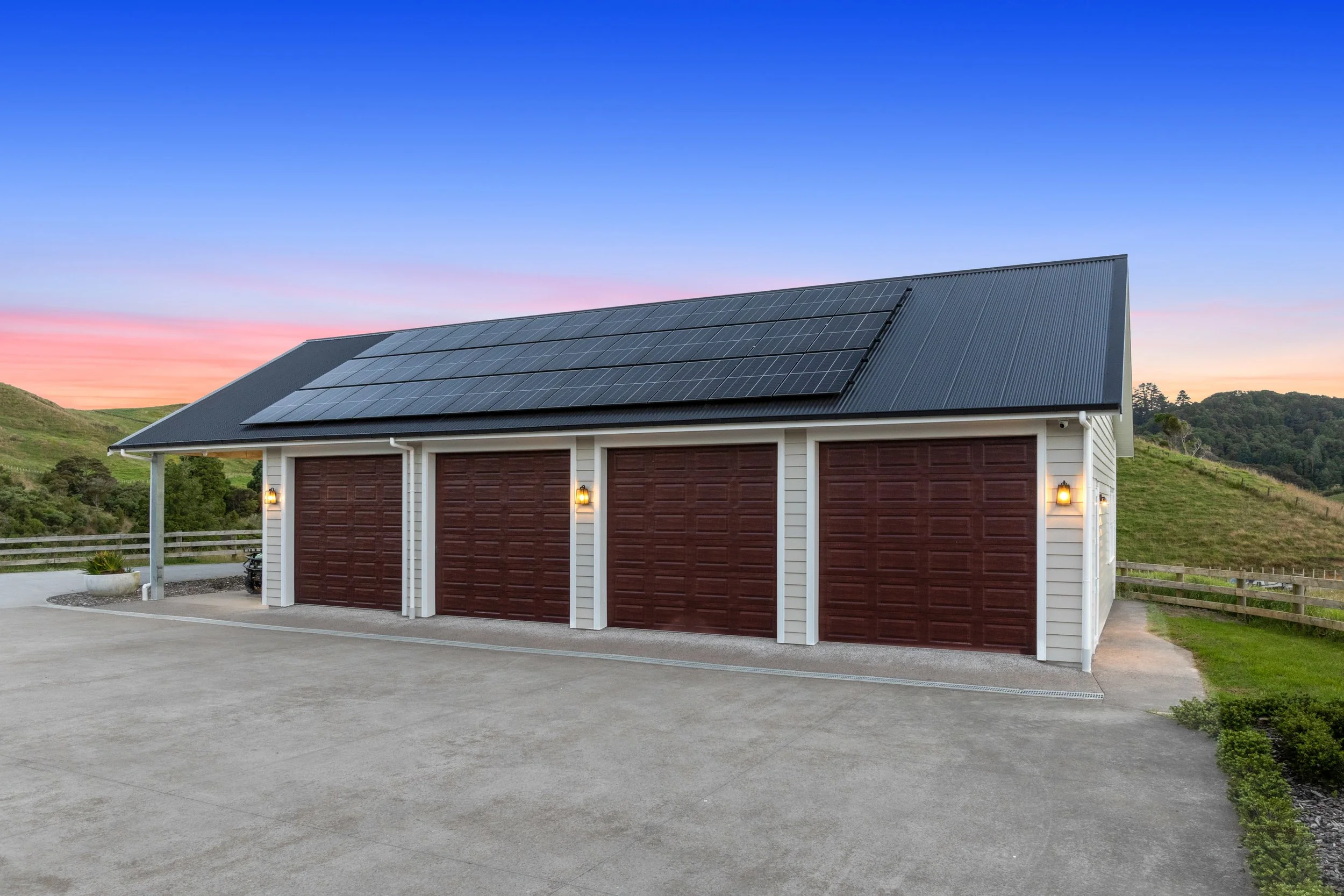 A residential garage with solar panels installed on the gray metal roof, featuring three garage doors with a wood-like finish, set against a landscape with green hills and a sunset sky.