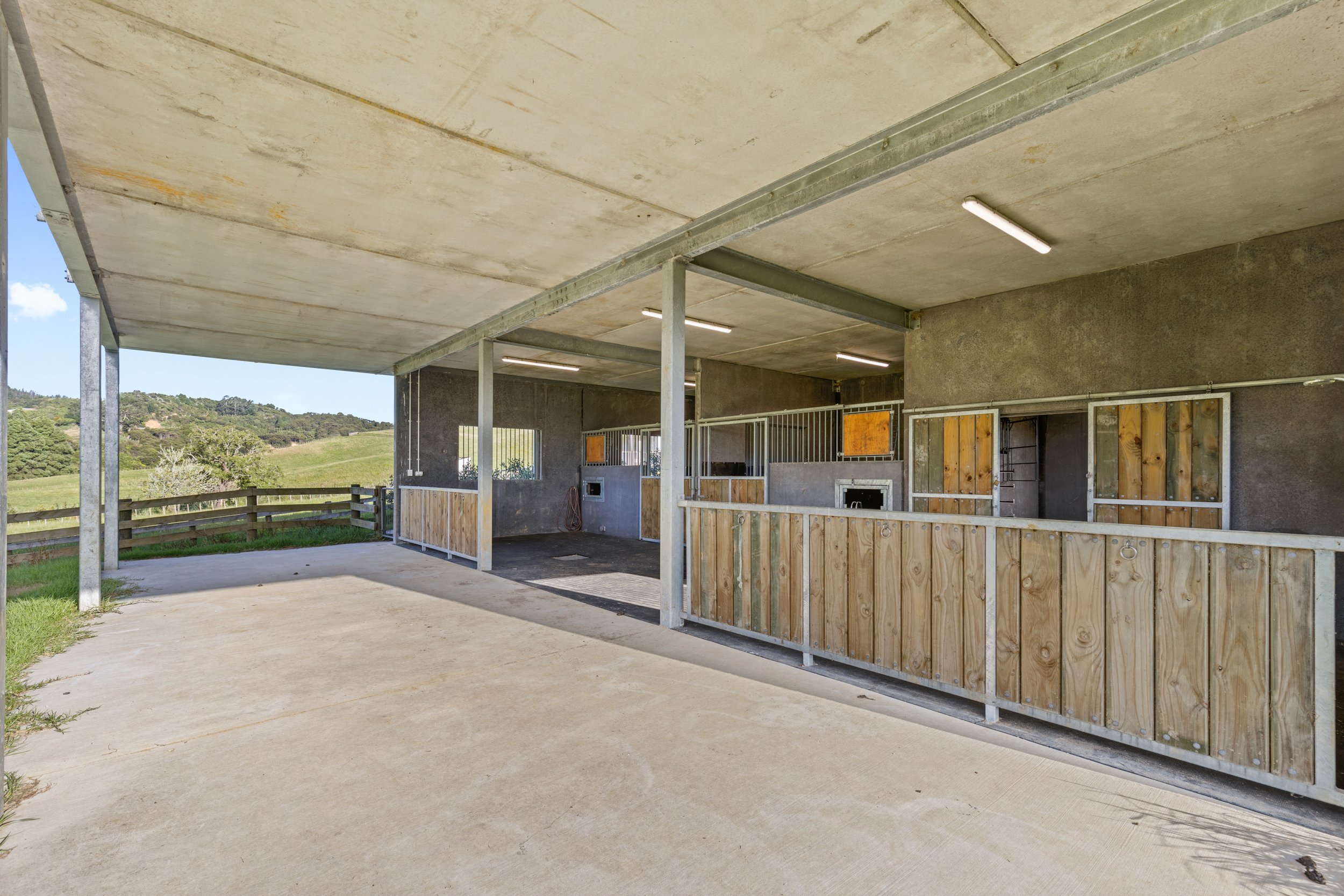 Concrete patio with a covered structure and fencing, overlooking a grassy landscape with hills and trees.