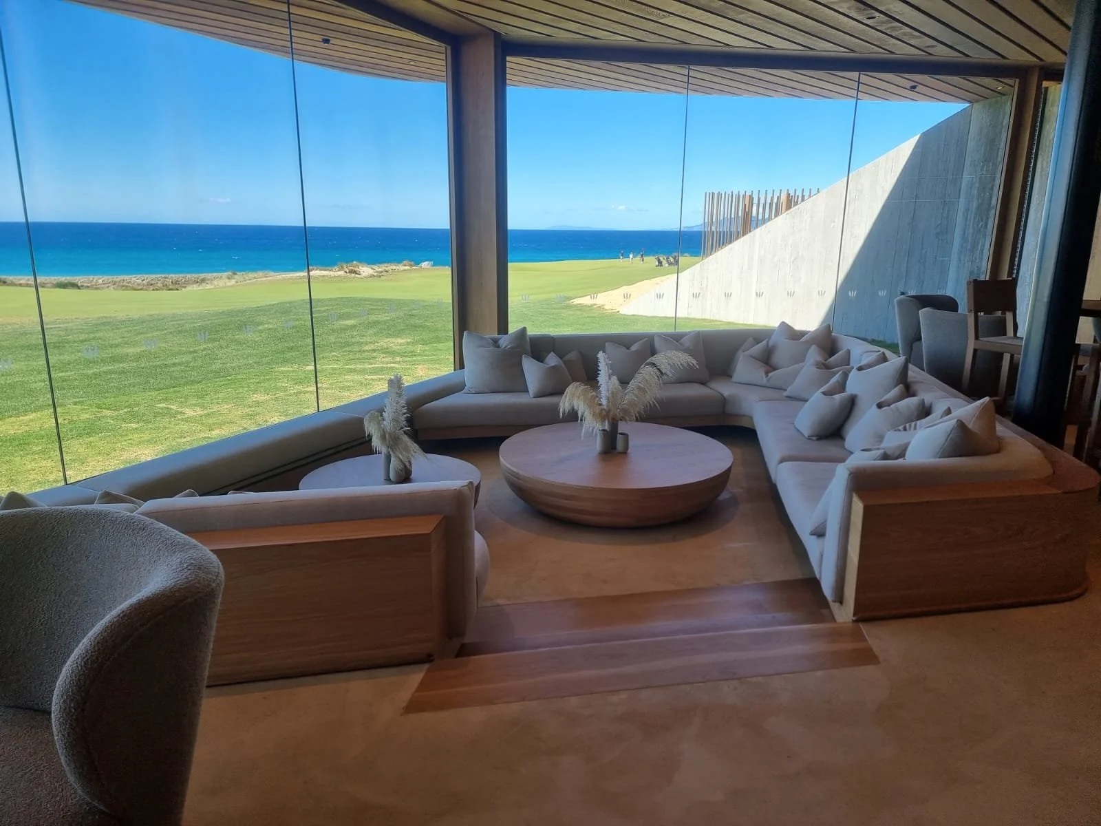 A cozy indoor seating area with a large U-shaped beige sofa and multiple white pillows, surrounded by a wooden coffee table with centerpieces of pampas grass, situated near a floor-to-ceiling glass wall that offers a scenic view of a grassy landscape