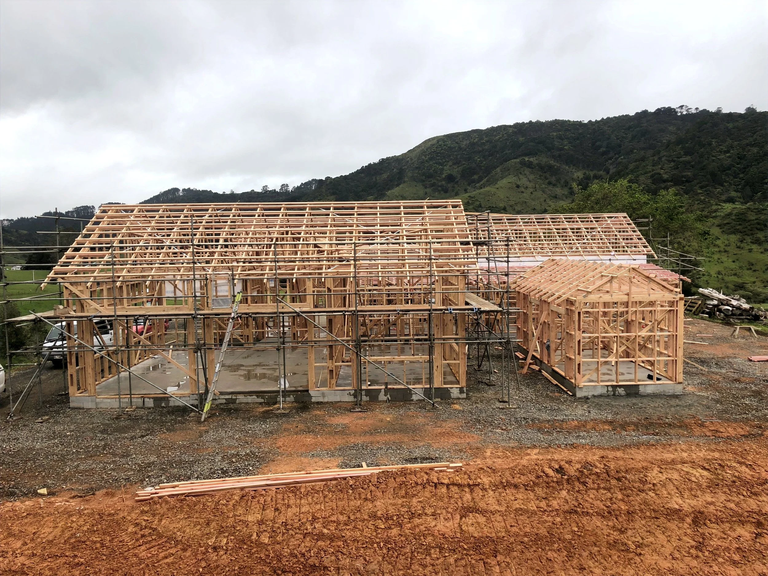 Wooden house construction site with scaffolding and framework in a rural, hilly landscape with cloudy sky.