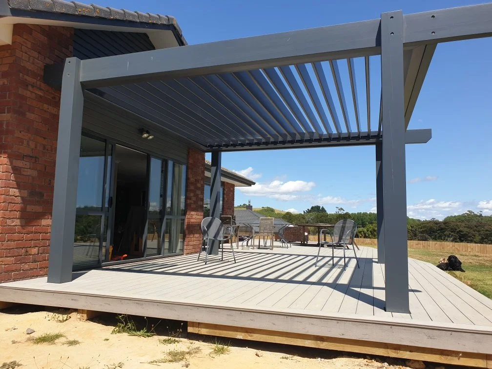 A backyard patio with a gray pergola, outdoor chairs, and a table, attached to a brick house, overlooking a scenic landscape with trees and blue skies.
